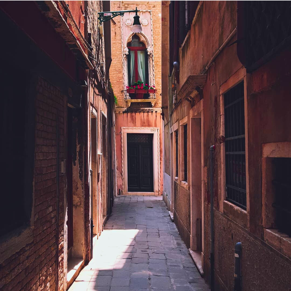 Sunlit narrow lane between brick buildings leading to an arched window with red flowers