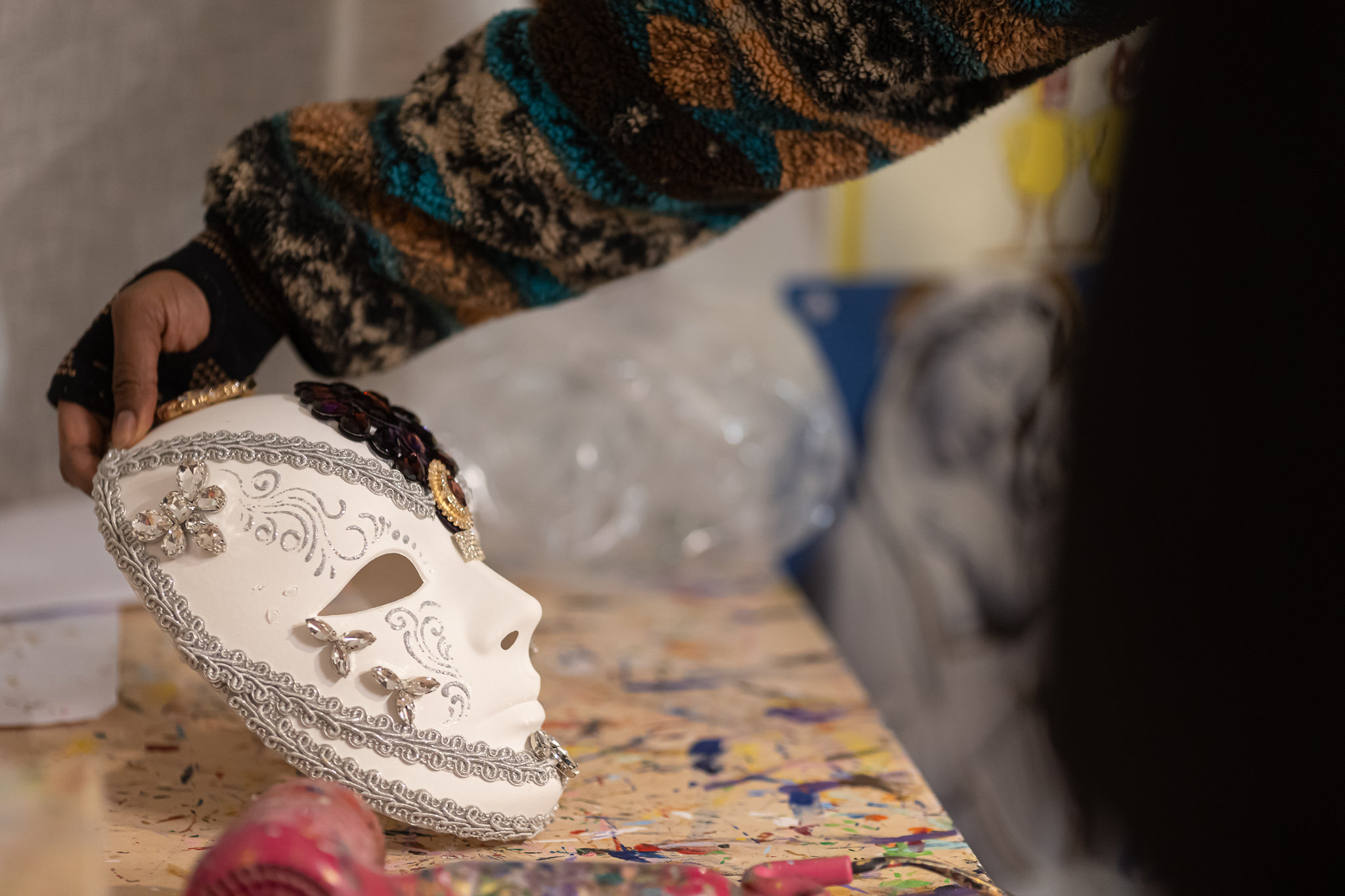 Hand holding a white Venetian mask decorated with silver lace and beads in a workshop