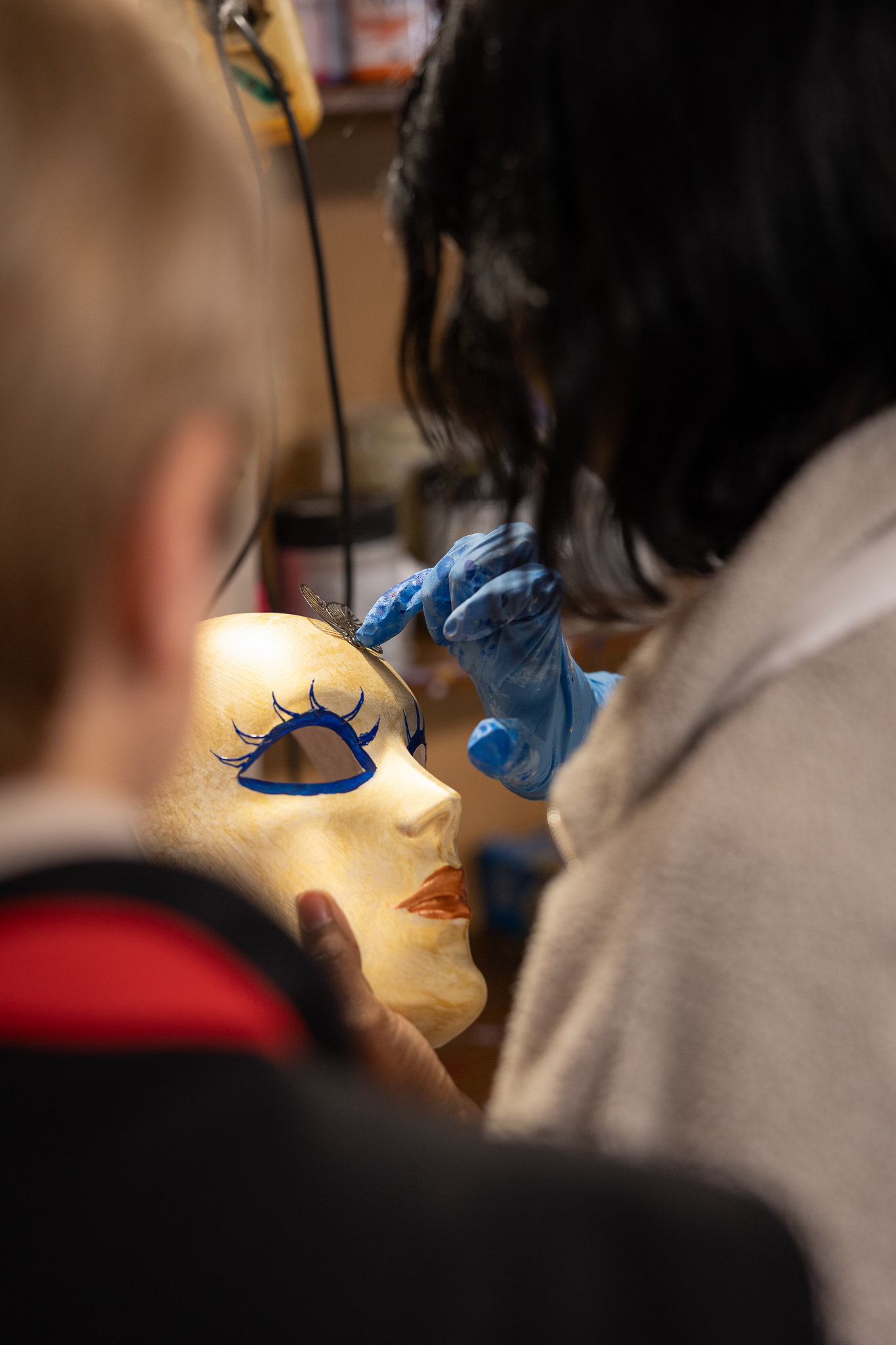 Artisan painting blue eye detail on a gilded mask while a visitor watches