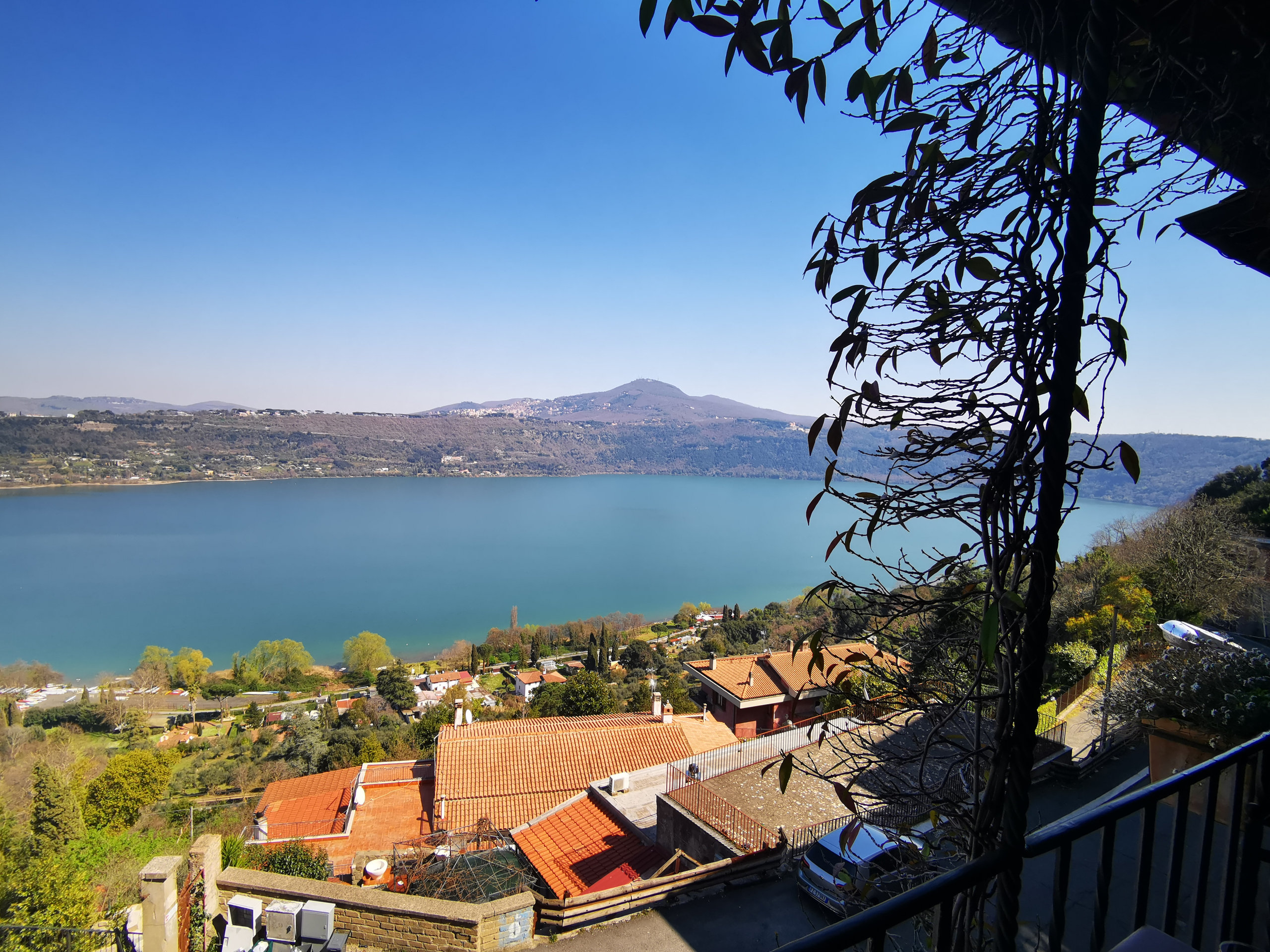 Blue crater lake seen from a terrace above terracotta rooftops, with distant hills