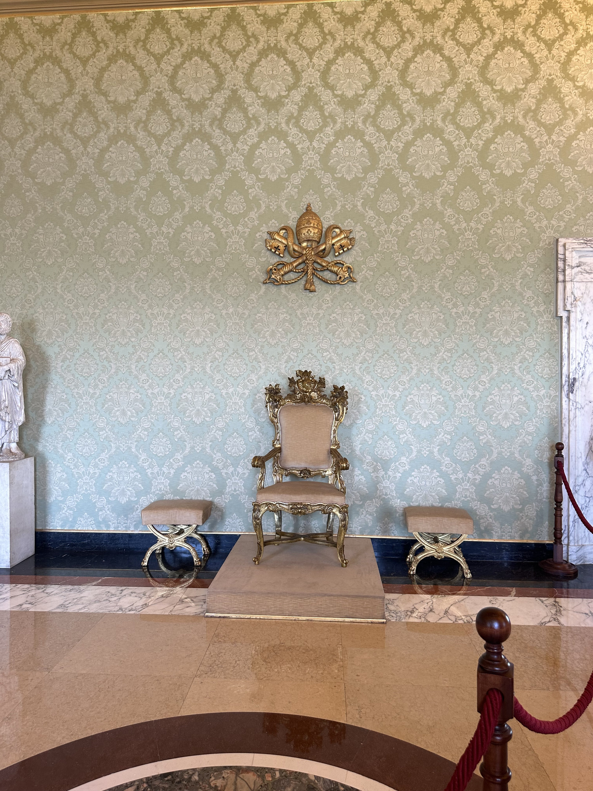 Gilded papal armchair and golden papal emblem against pale green damask walls