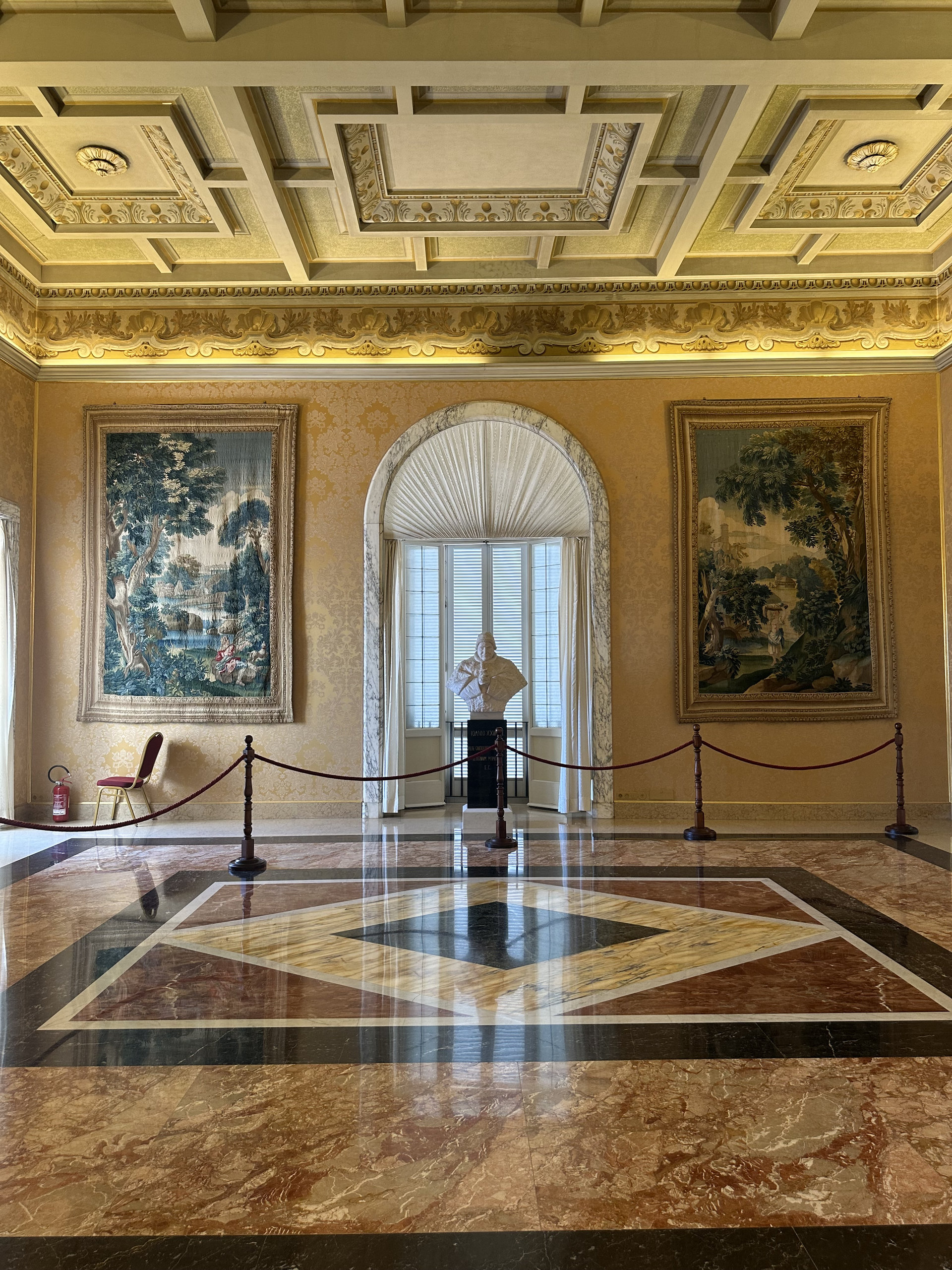 Palace hall with tapestries, a marble bust and a geometric marble floor behind velvet ropes