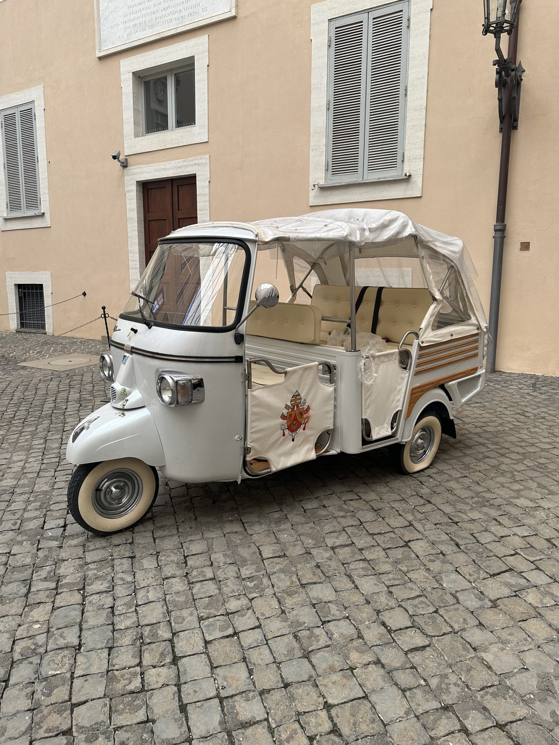 Cream-coloured three-wheeler tuk-tuk with a papal crest parked on a cobbled courtyard