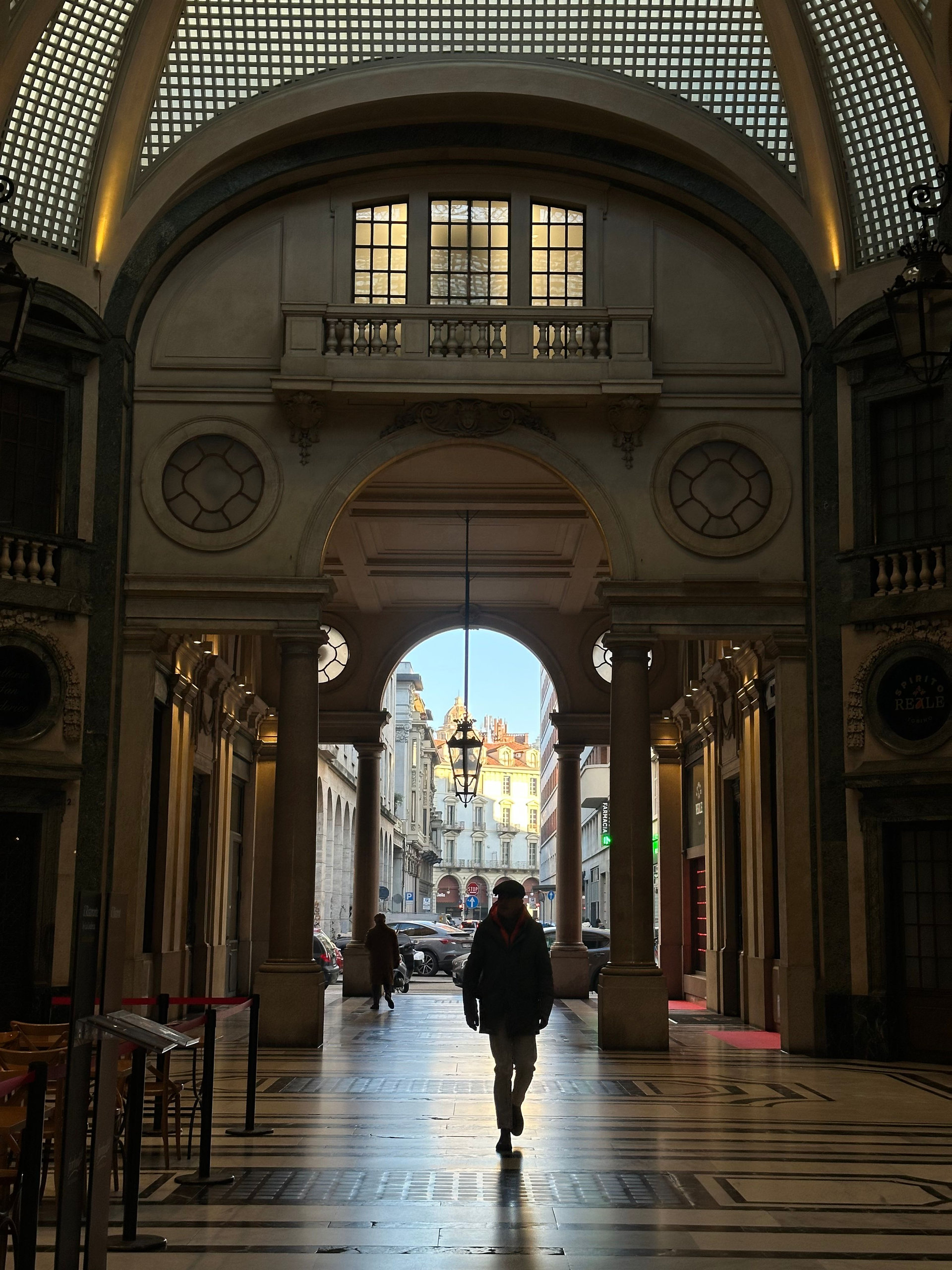 Walker crossing an arcaded passage with a glazed barrel vault leading to a sunlit street in Turin