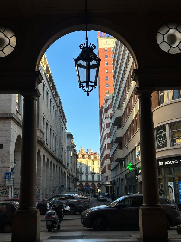 View through an arcade arch with a wrought-iron lantern onto a Turin street and tall buildings