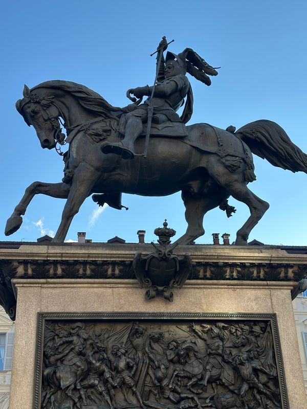 Bronze equestrian monument with a battle relief on its pedestal, seen from below against blue sky