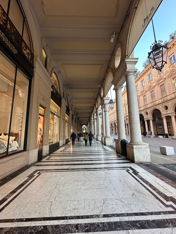 Long marble-floored arcade with shops and lanterns leading to a sunlit square