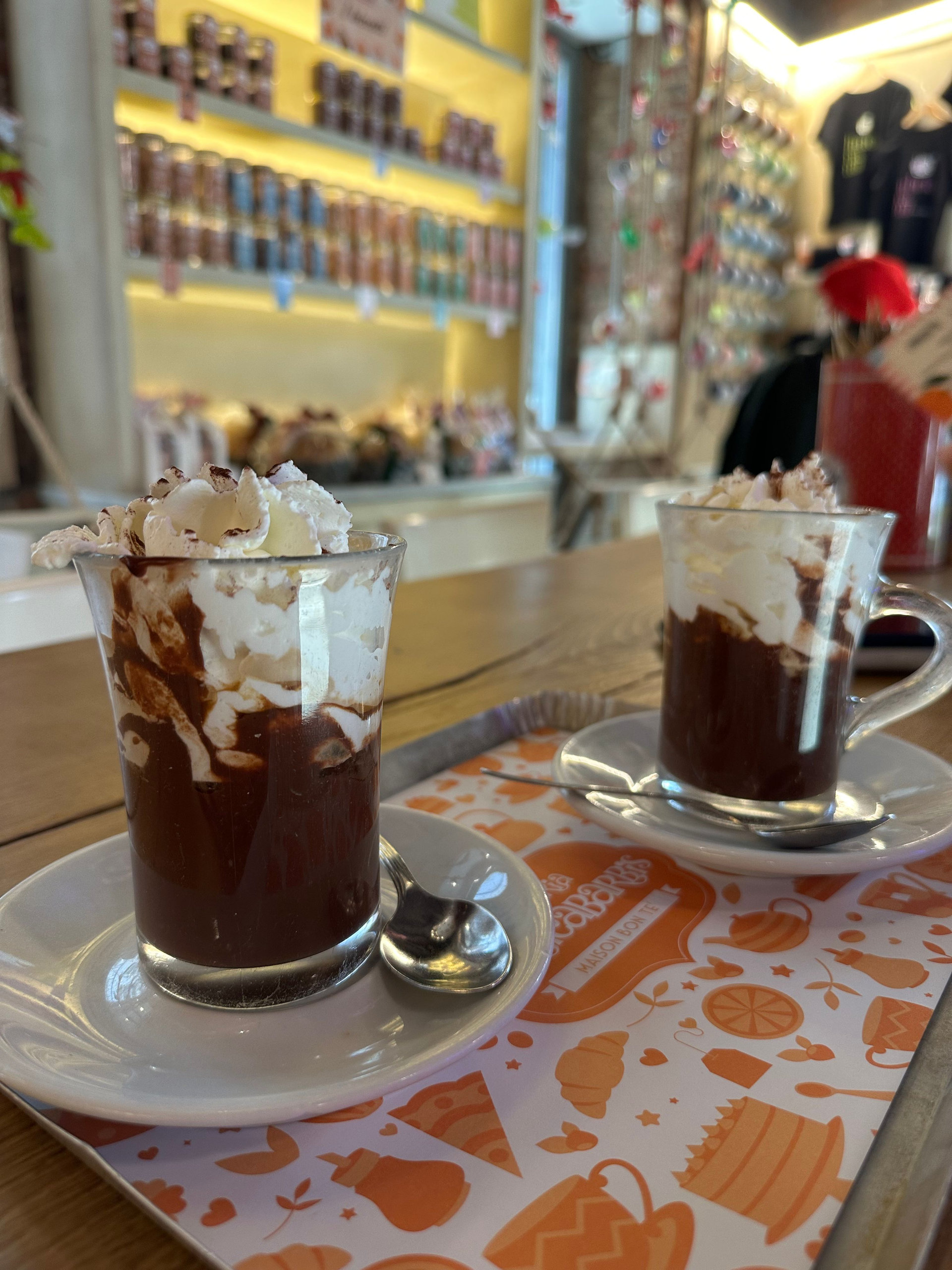 Two glasses of hot chocolate topped with whipped cream on a patterned tray at a café counter