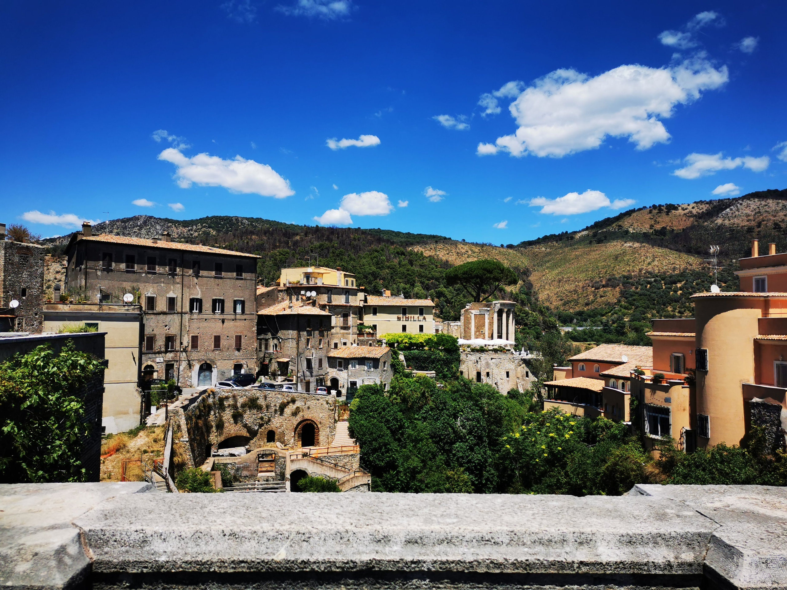 Tivoli rooftops and a hilltop round temple seen over a stone parapet beneath a deep blue sky