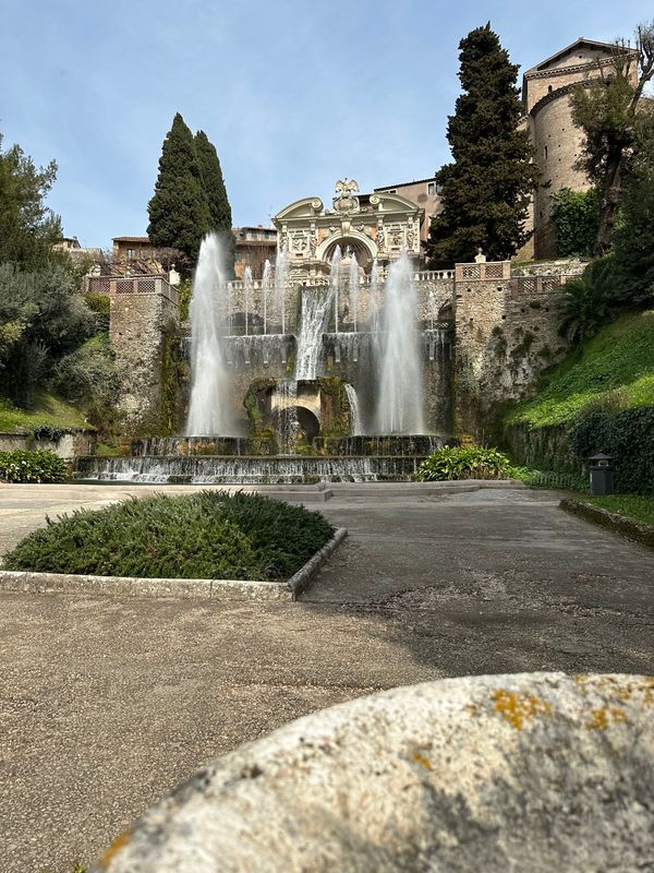Fountain with jets of water below a carved stone pavilion, framed by cypress trees and a pathway