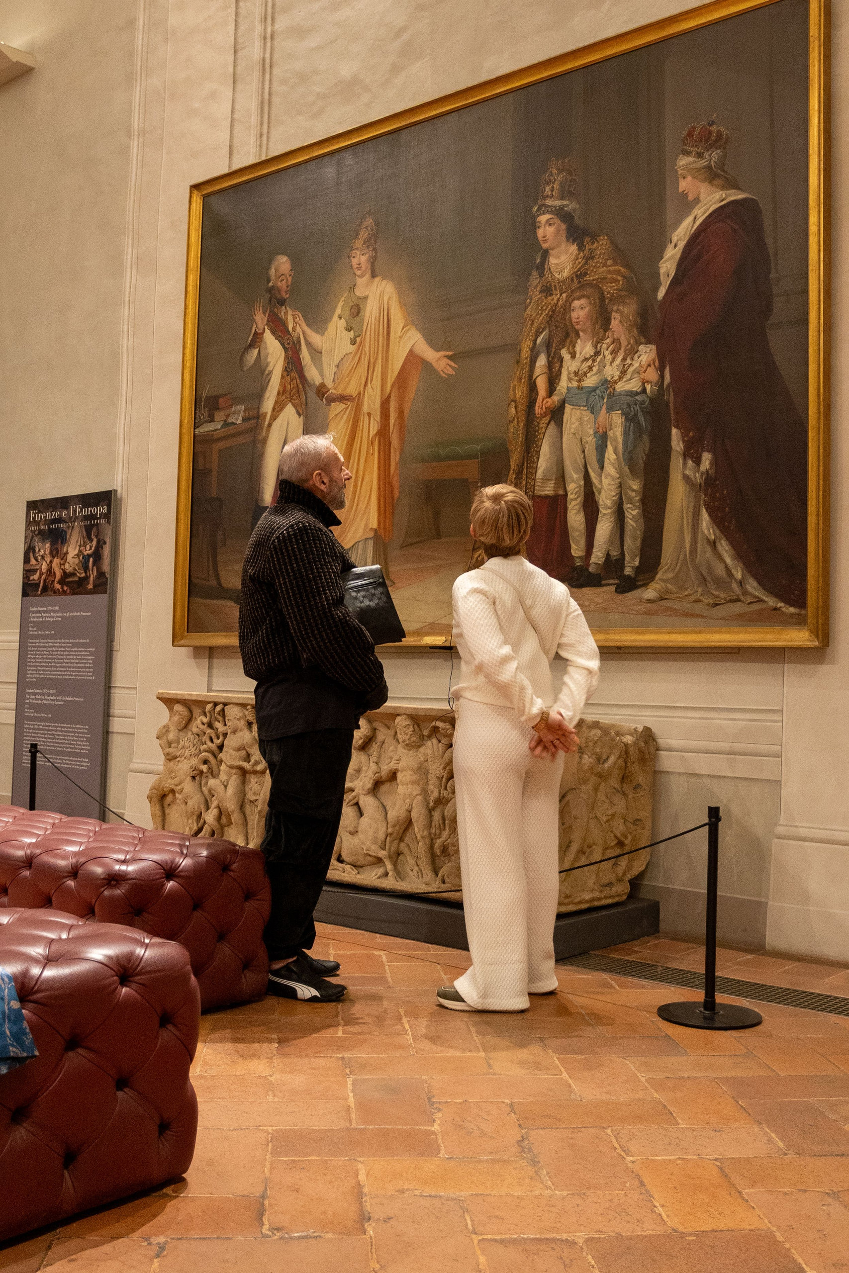 Two visitors admiring a large royal portrait in a museum hall with leather benches