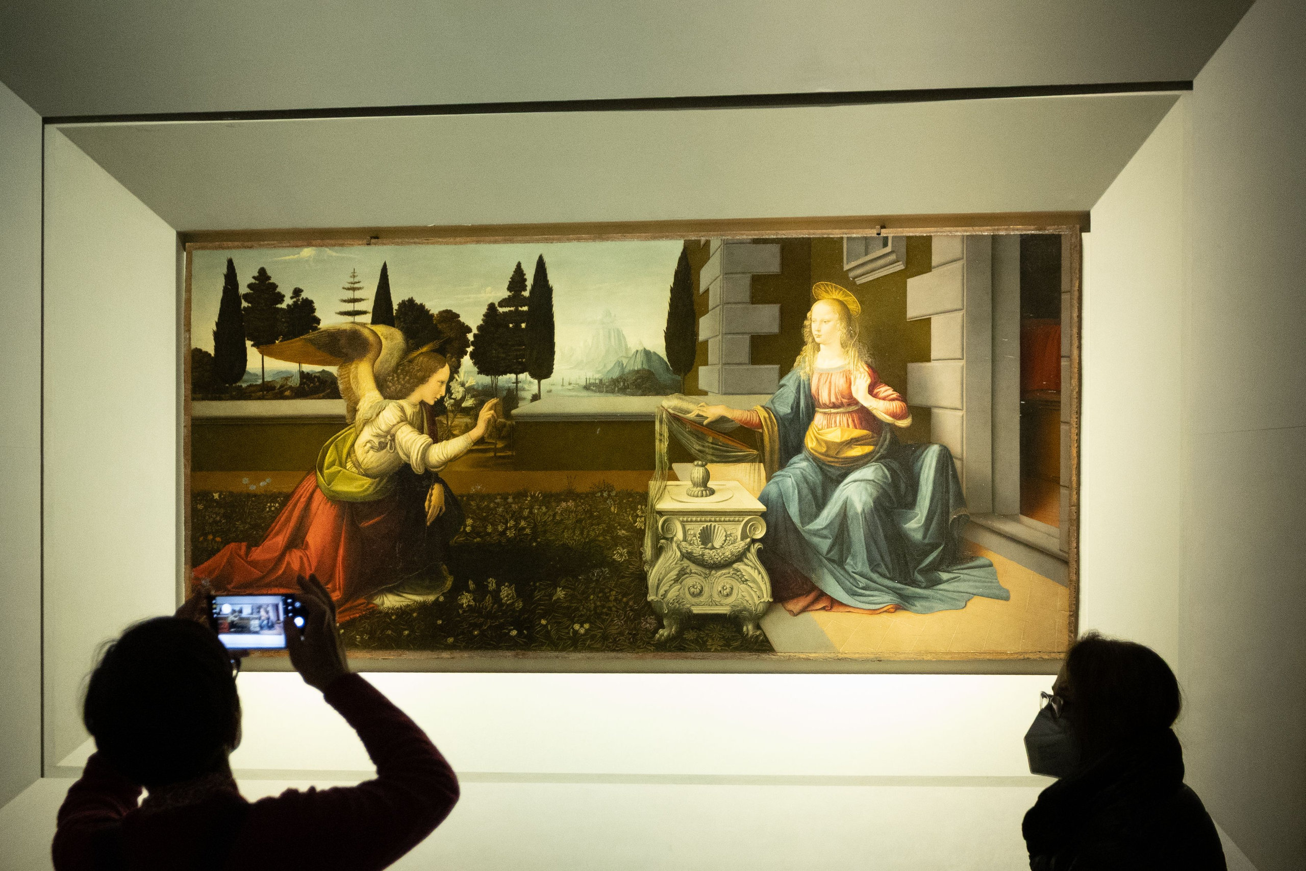 Visitors photographing a Renaissance Annunciation painting in a dimly lit gallery room