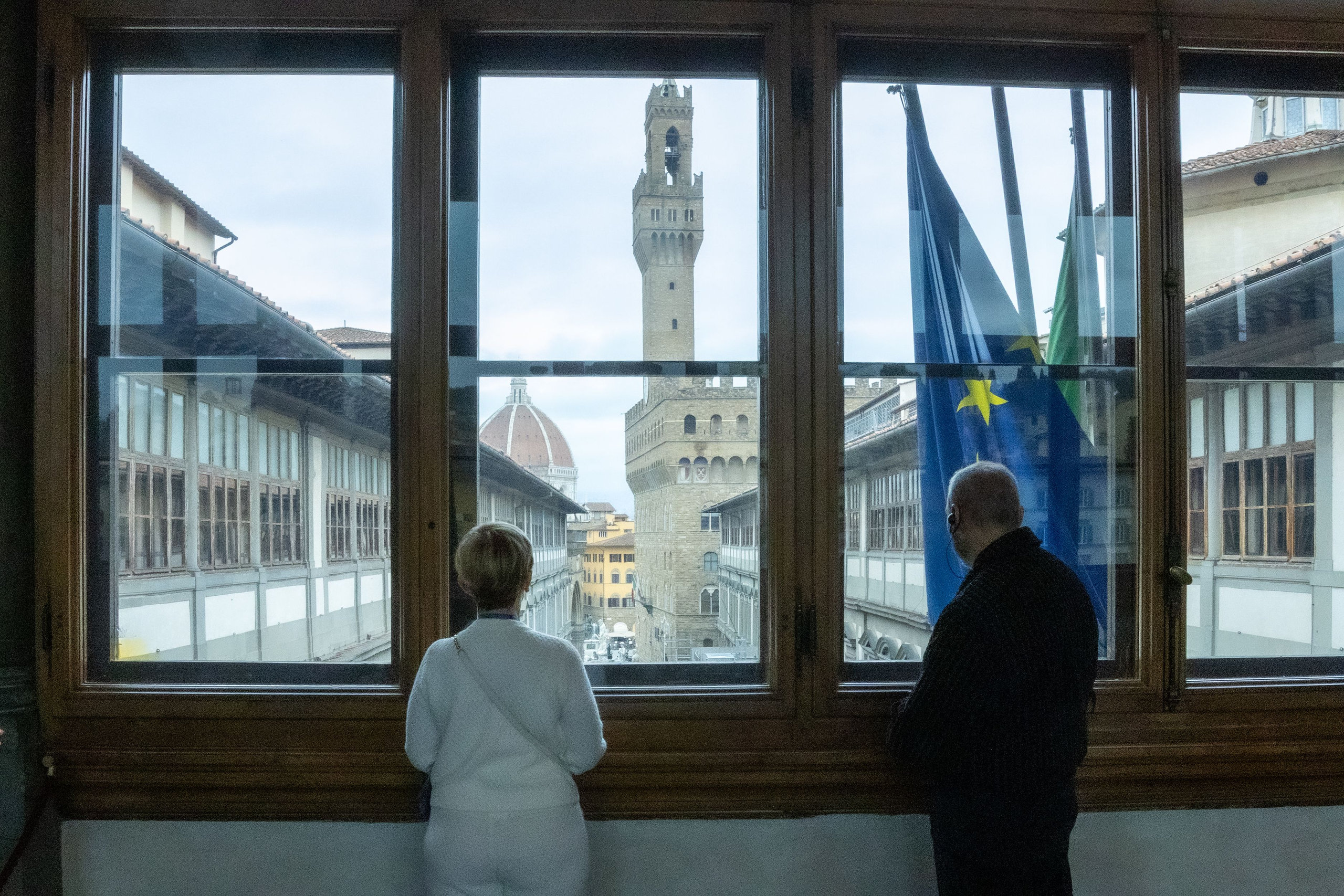 Two travellers looking through a gallery window at the Palazzo Vecchio tower and an EU flag