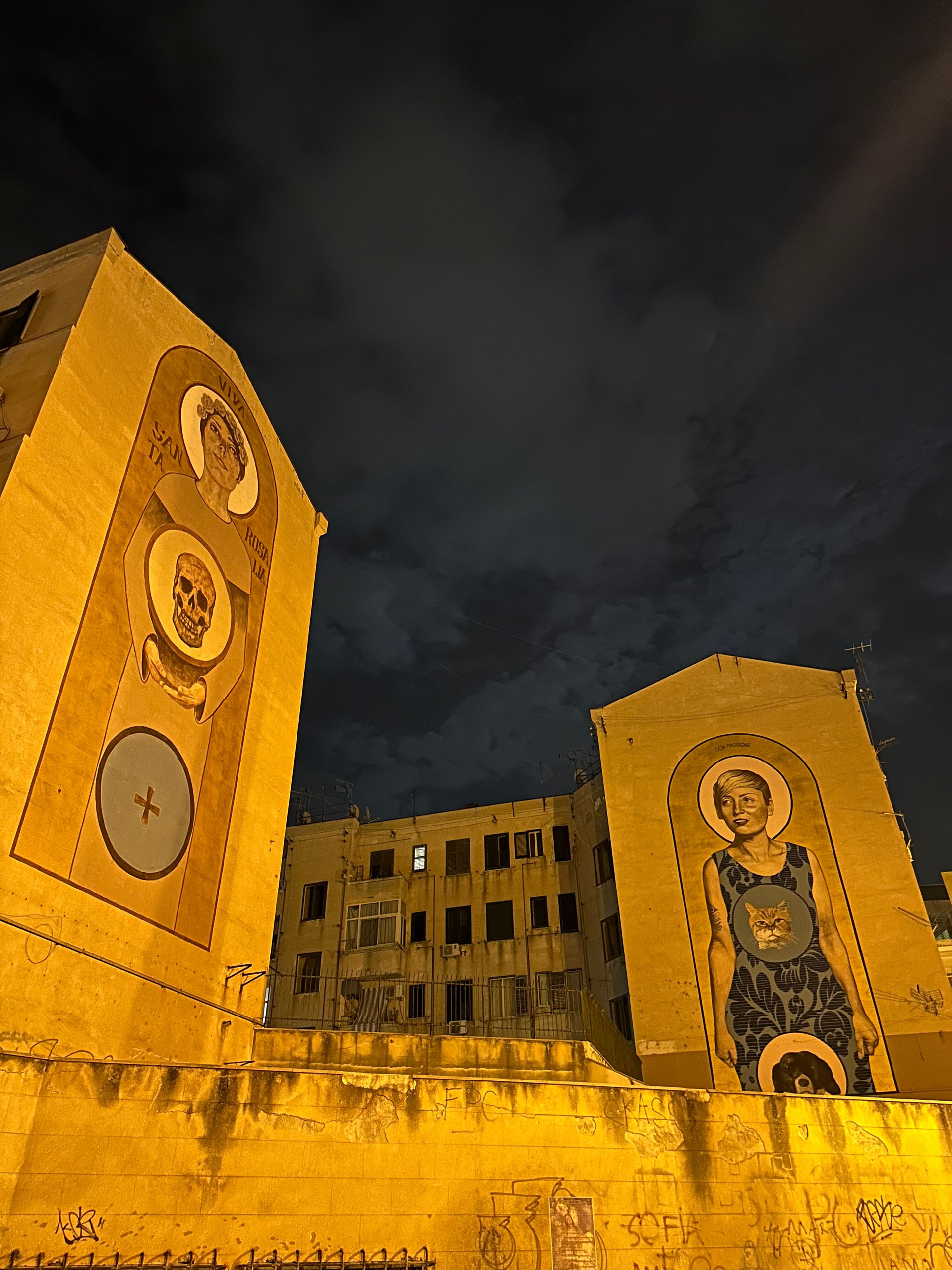 Large illuminated murals of portraits on building walls against a dark night sky