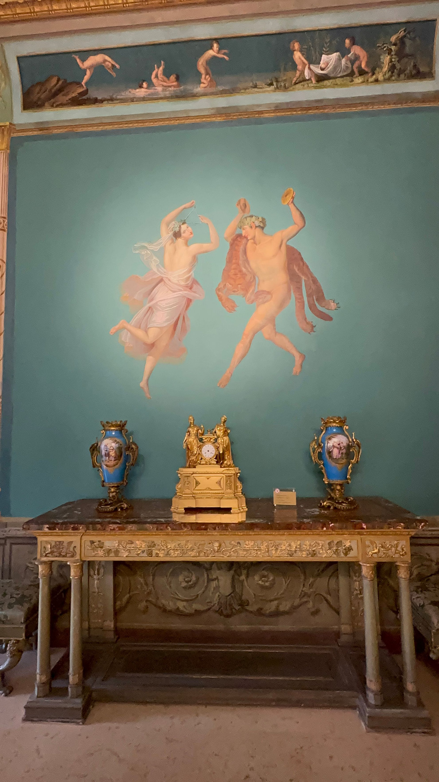 Painted wall with two dancing figures above a gilded console, clock and blue vases