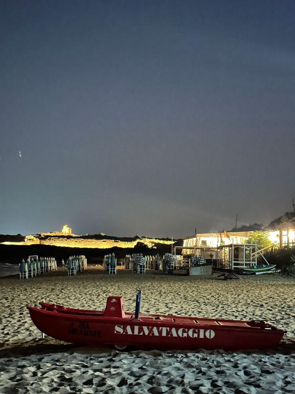 Red lifeguard boat on a sandy beach at night with beach huts and lights along the shore