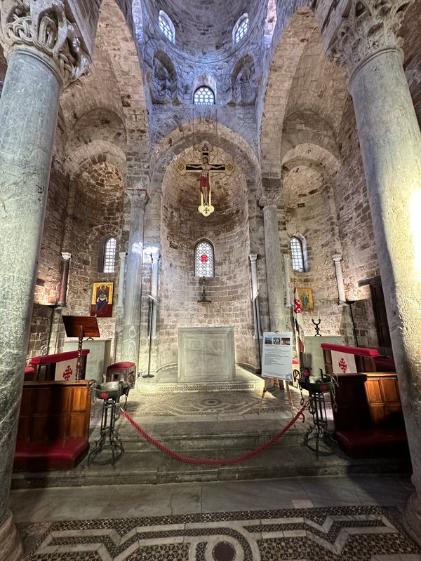 Candlelit Norman-Byzantine church interior with stone columns, a hanging crucifix and a mosaic-tiled floor