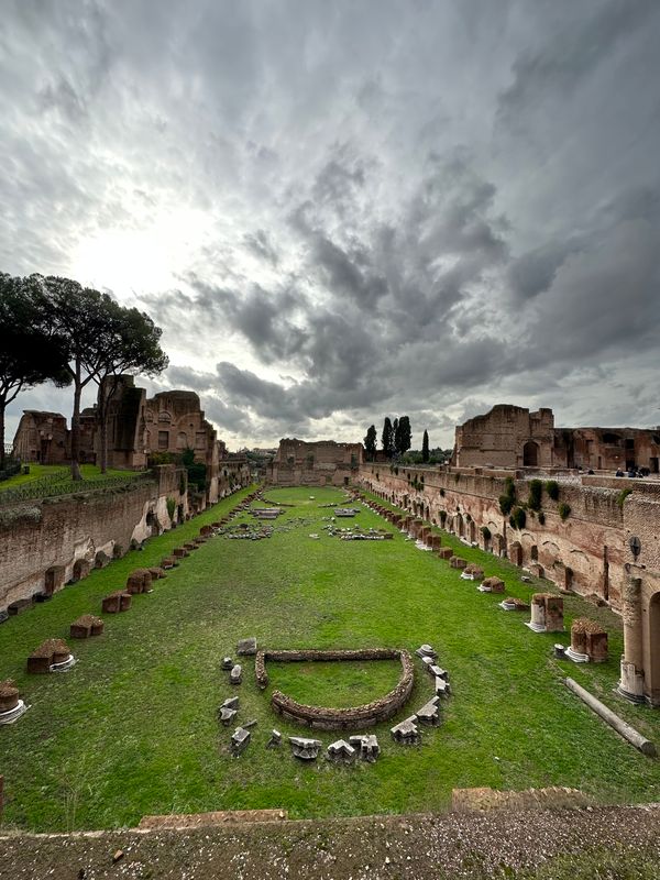 Grassy ancient stadium on the Palatine Hill with ruined brick arcades under a stormy sky
