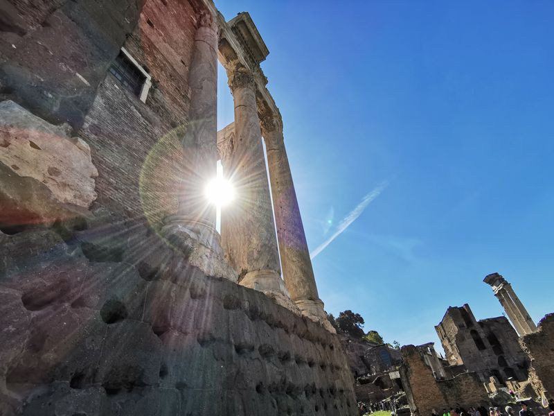 Sun flaring behind ancient Roman columns rising from weathered brick walls in the Forum