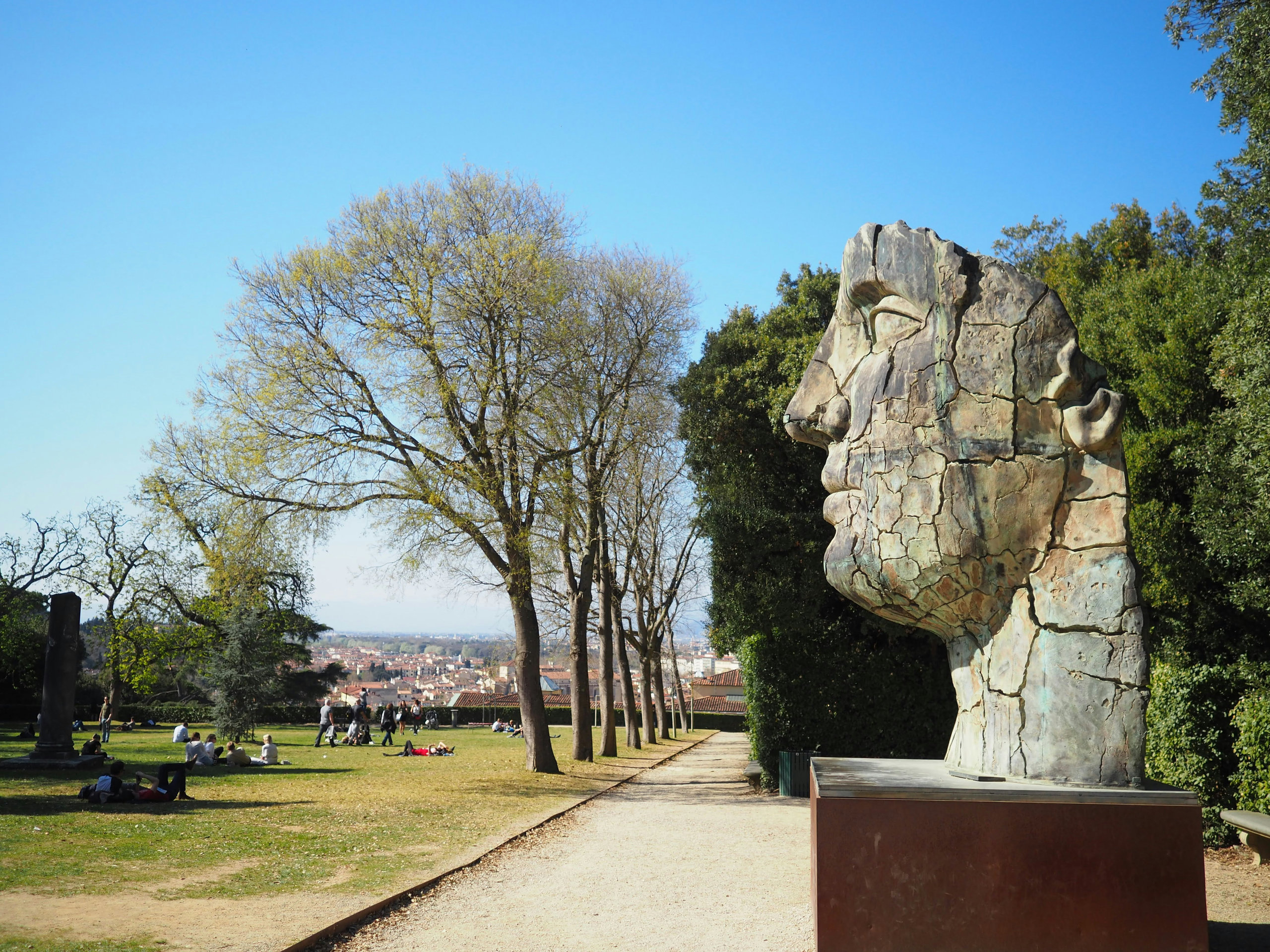 Large stone head sculpture beside a tree-lined path in the Boboli Gardens, with a view over Florence