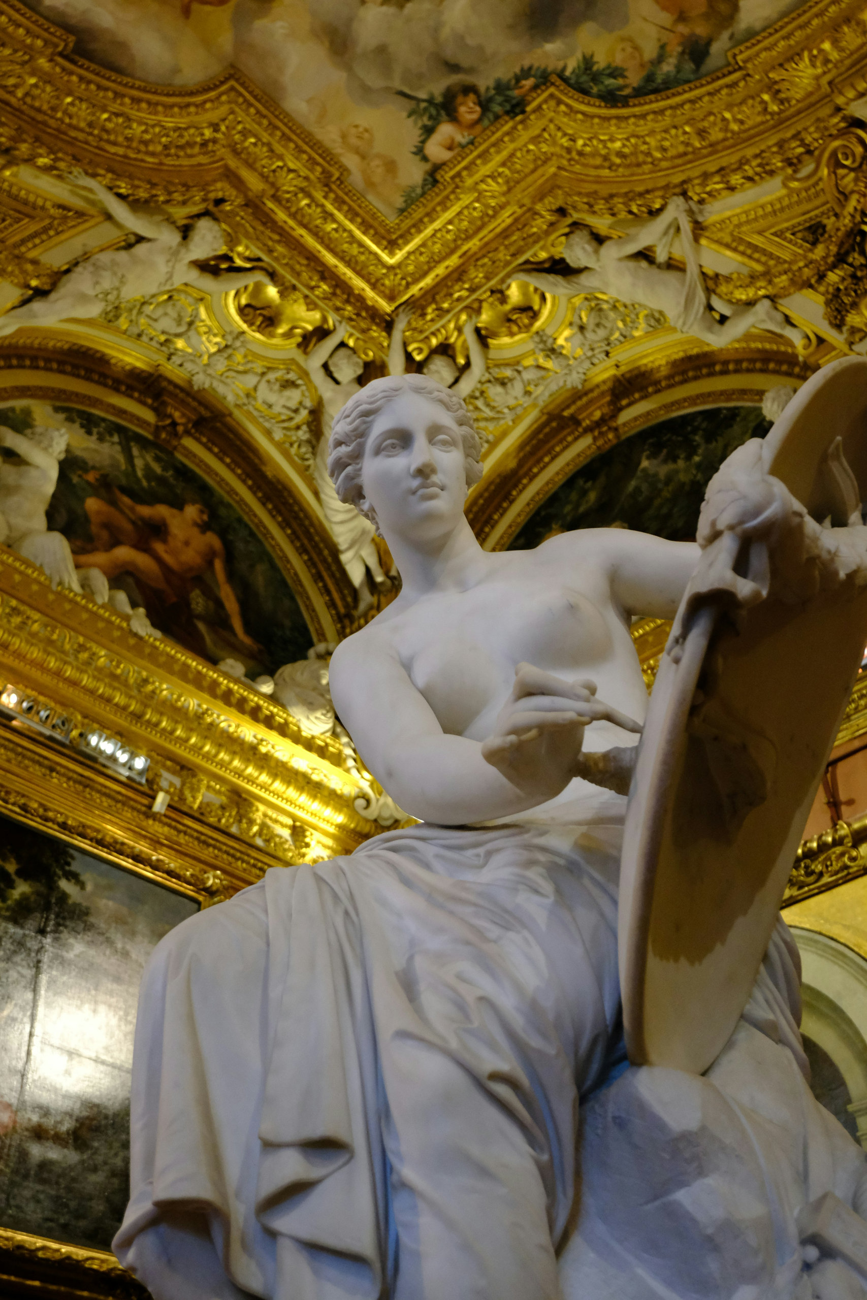Marble statue of a seated woman holding a palette beneath a gilded, frescoed ceiling