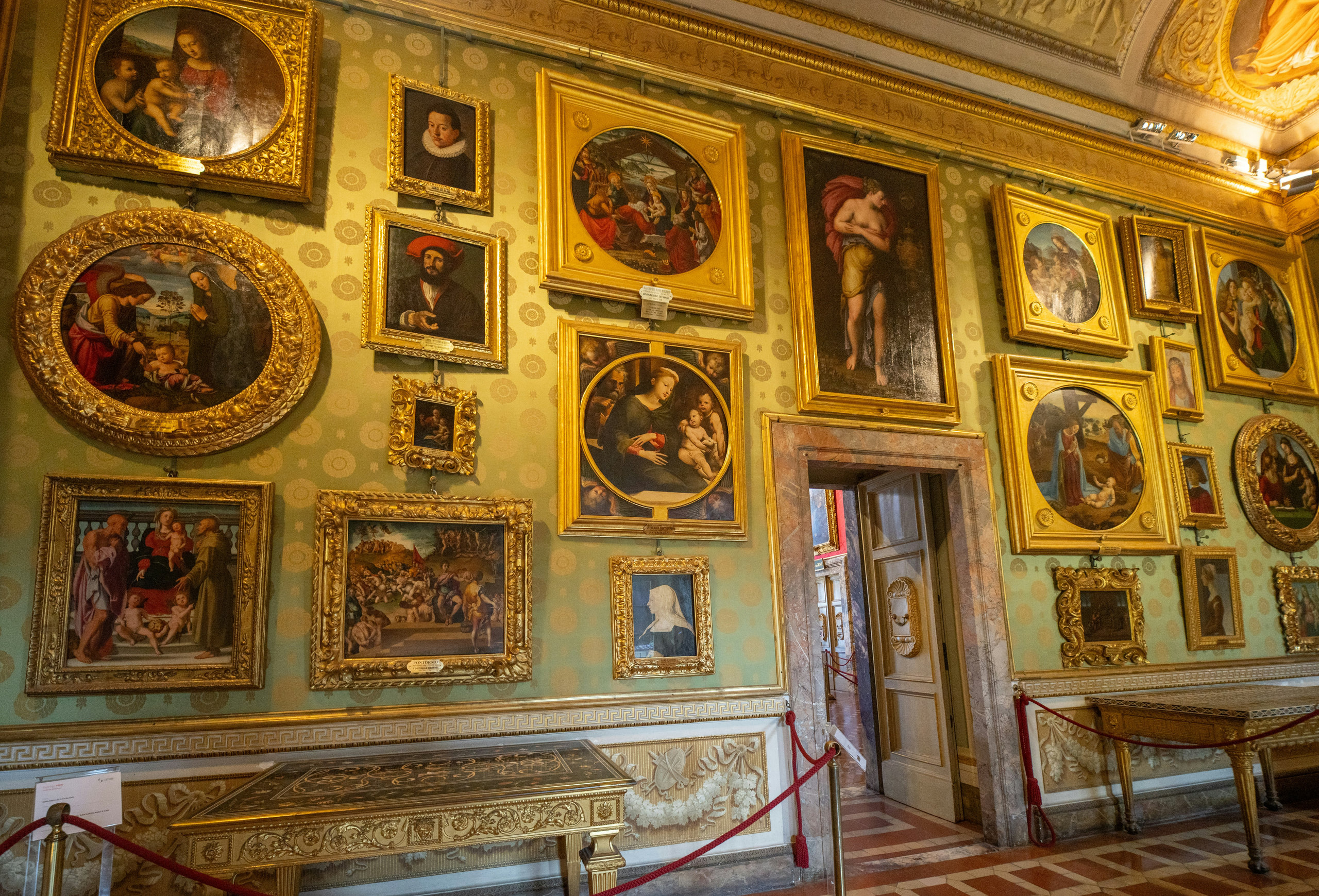 Salon wall densely hung with gilt-framed Renaissance paintings above a red velvet rope