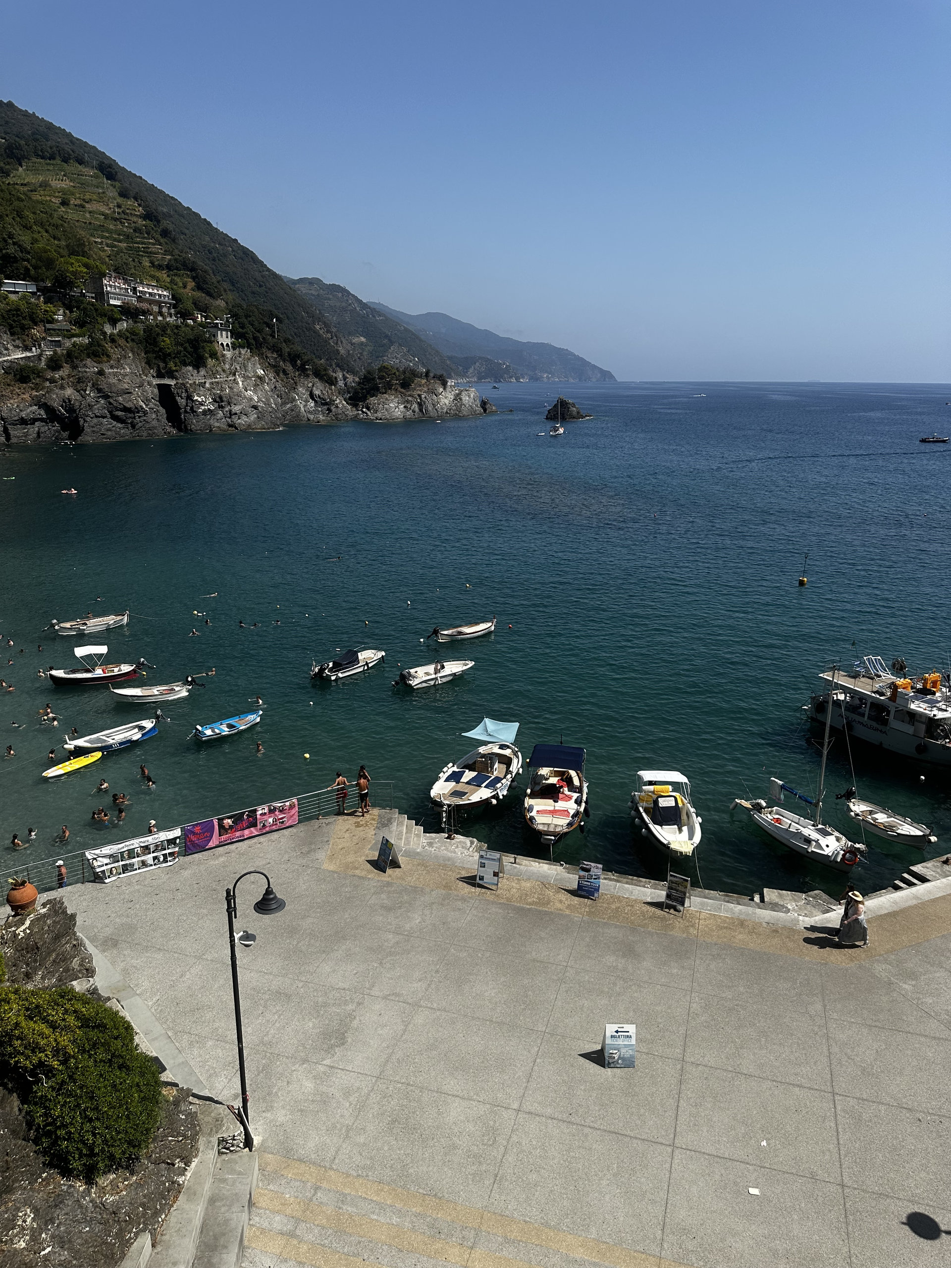 Small boats moored in a deep blue bay below a paved terrace, with green headlands in the distance