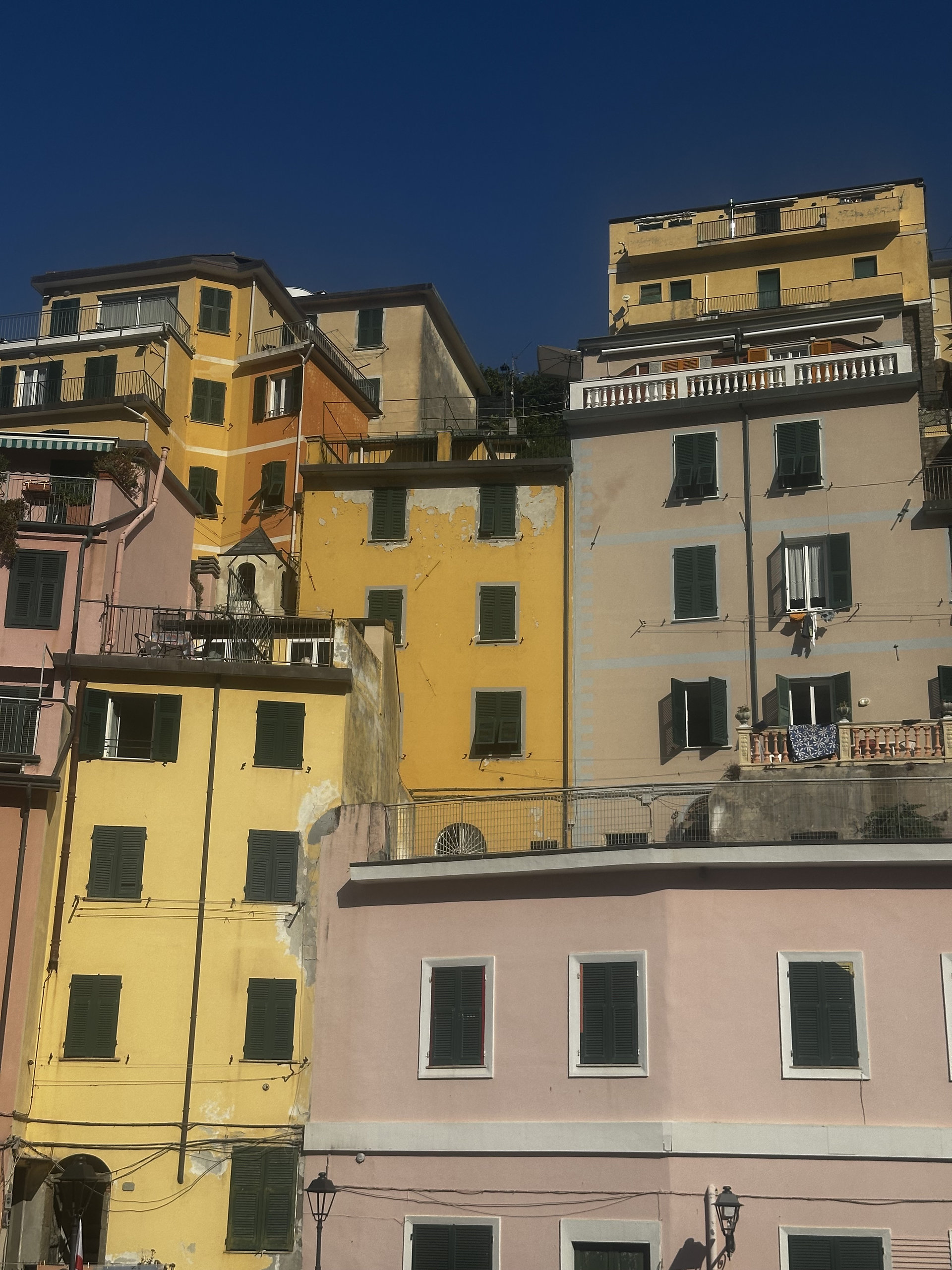 Tall ochre, yellow and pink houses with green shutters stacked up a hillside under a blue sky