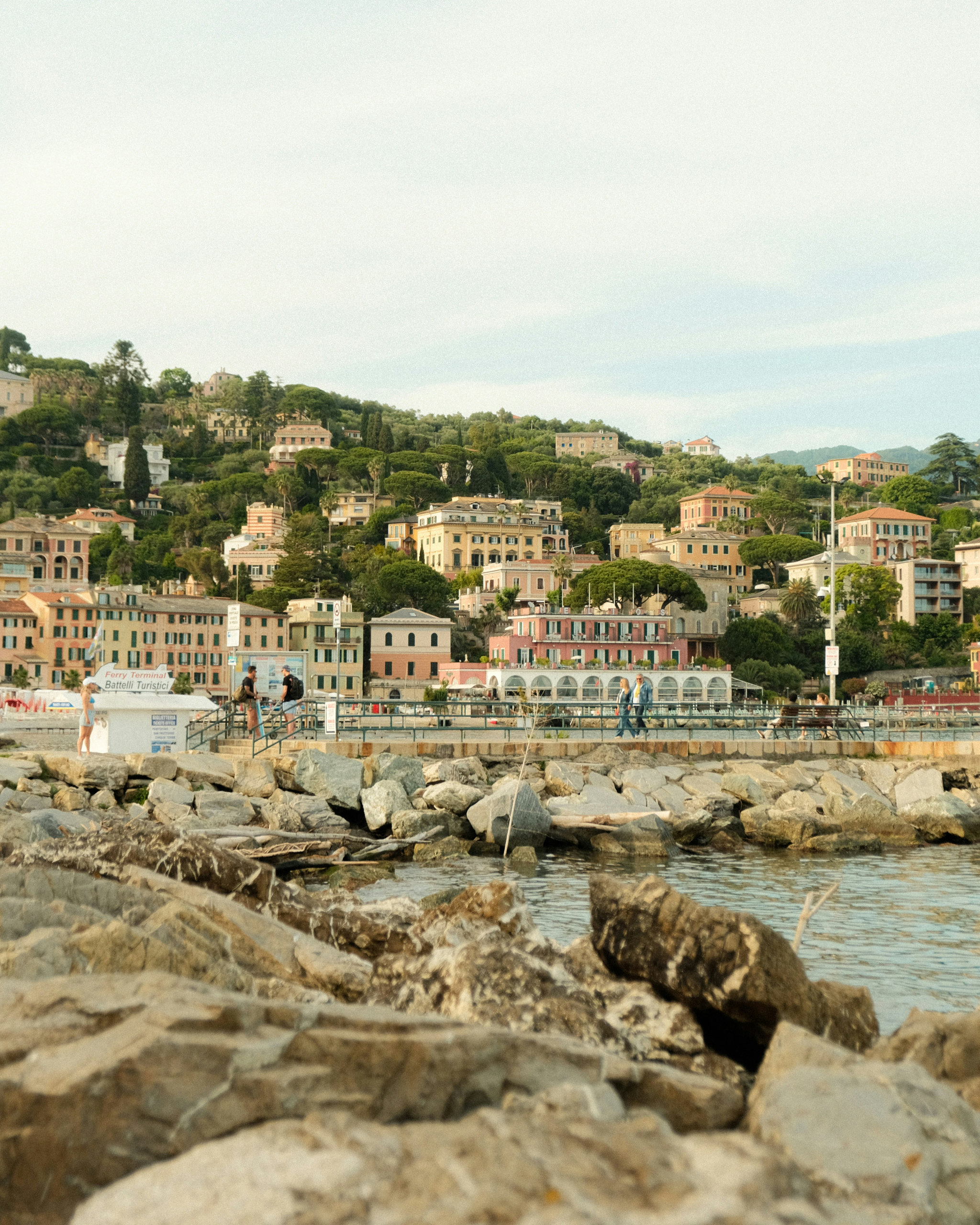 Pastel seaside town on a wooded hillside seen across rocks on the shore