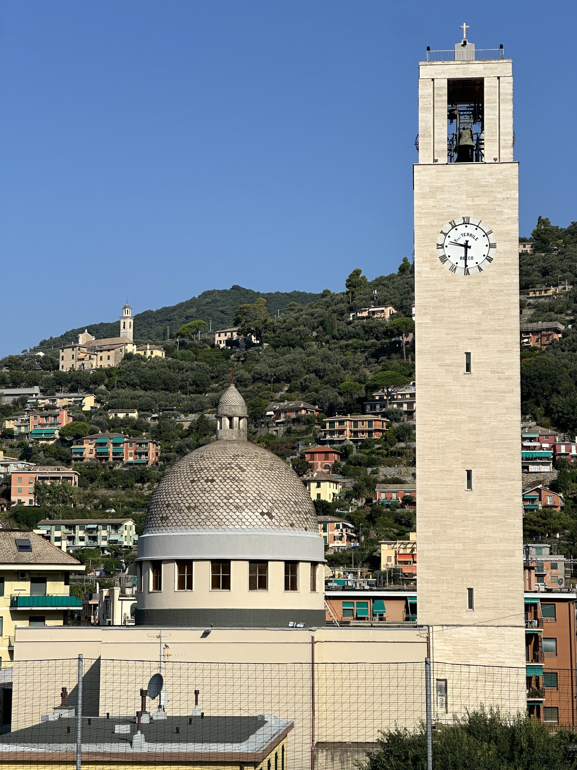 Domed church beside a tall clock tower, with coastal houses climbing the green hills behind