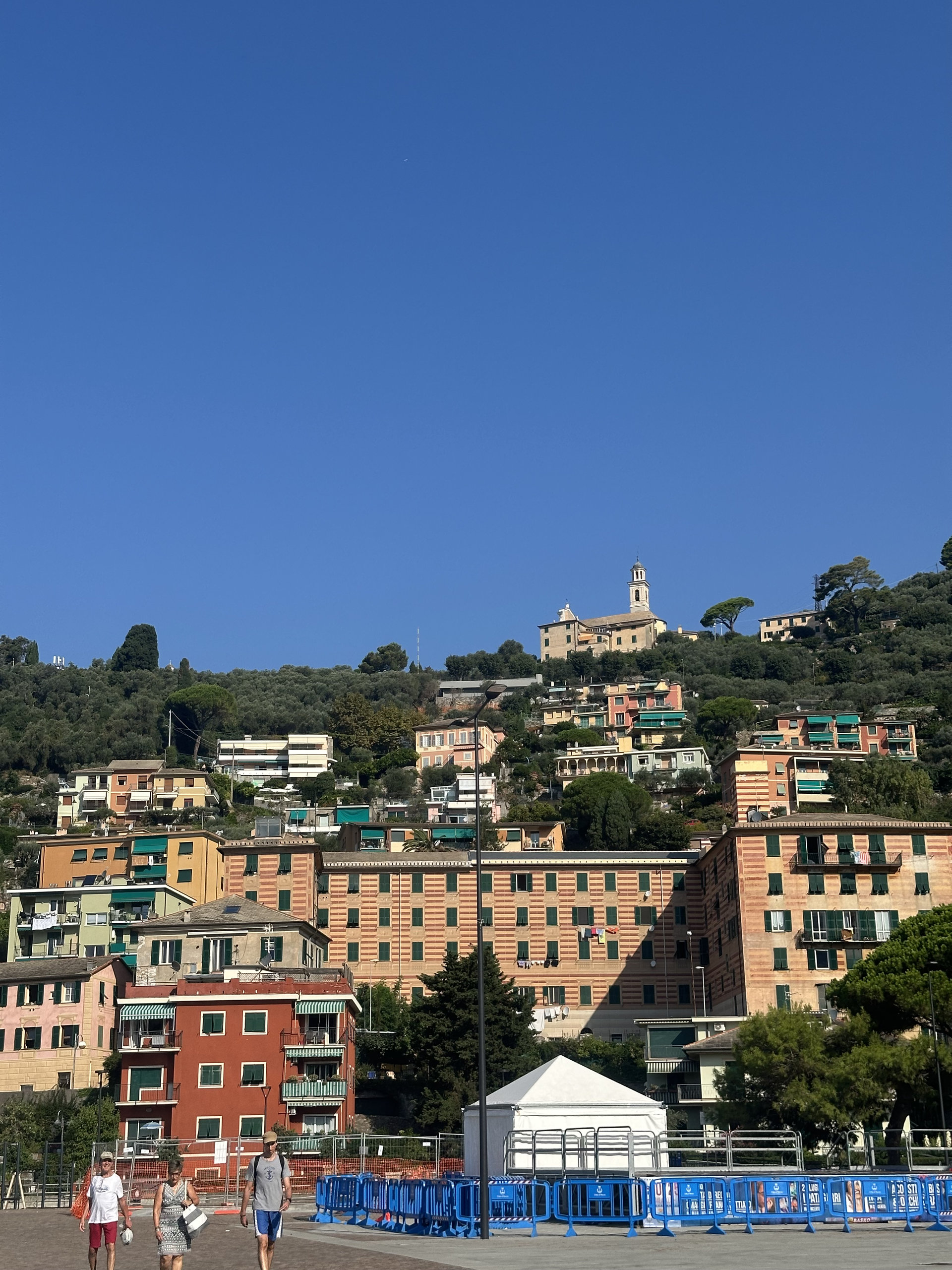 Pastel apartment buildings on a hillside above a seafront square under a clear blue sky