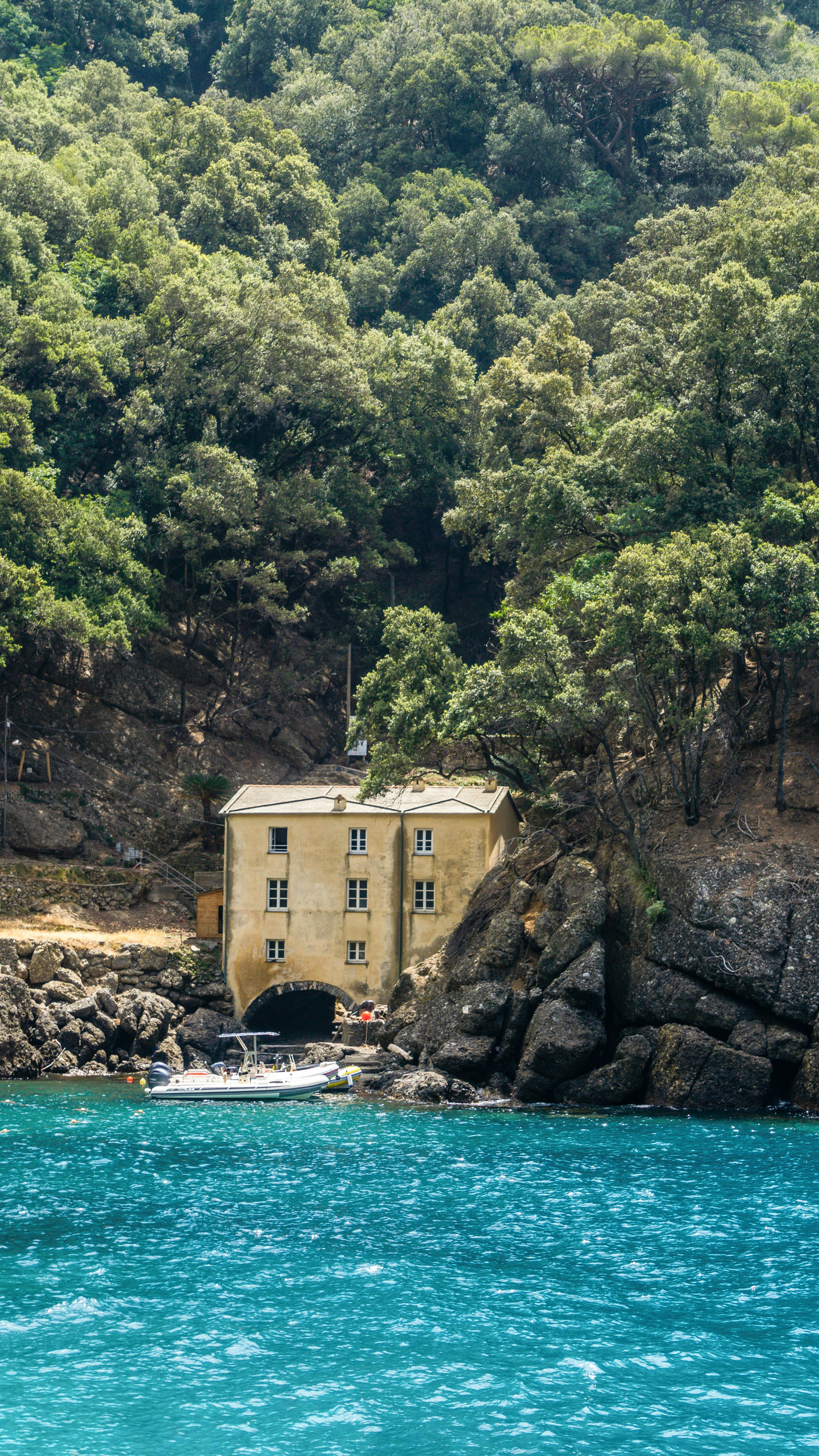 Ochre house with a boat at its arched landing on a wooded rocky cove above turquoise water