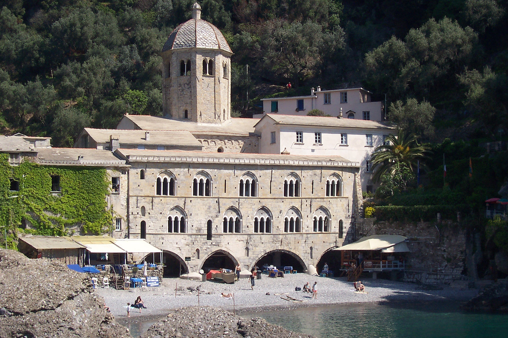 Medieval abbey with arcaded loggia and bell tower on a small beach, backed by dark green woods