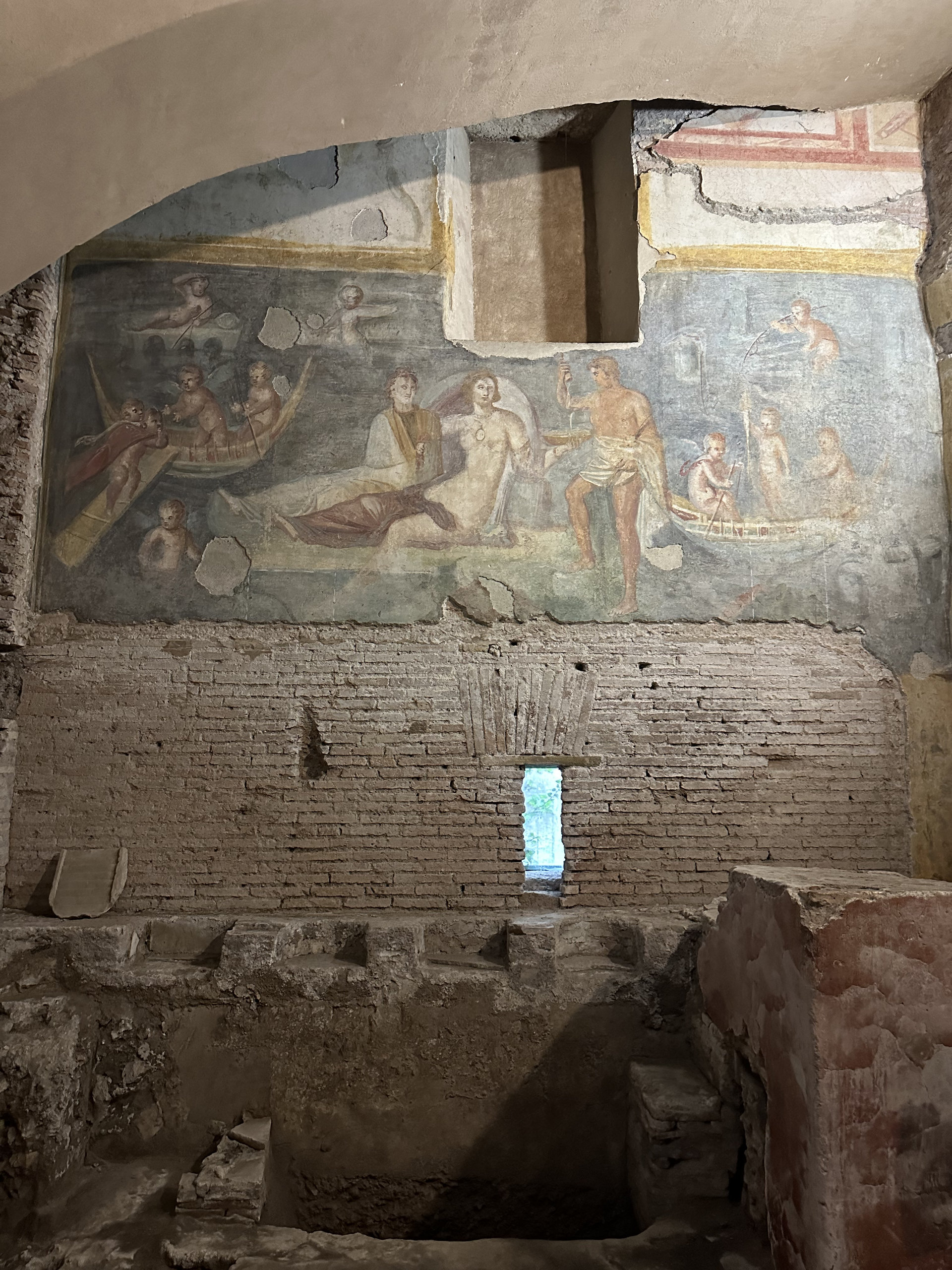 Chamber with a colourful fresco of boats and figures above weathered brick walls and a red masonry block