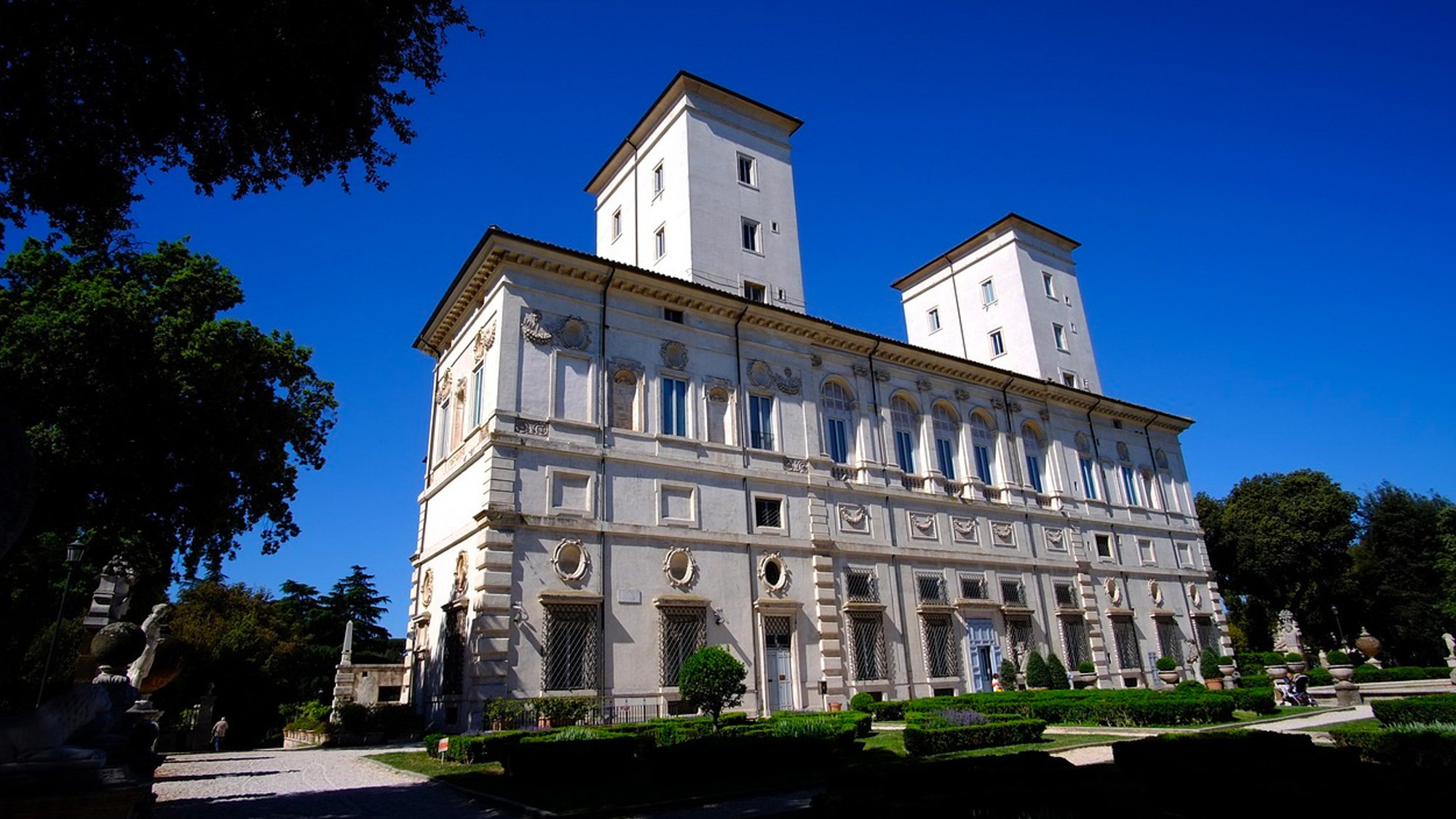 Cream-coloured Borghese Gallery villa with twin towers under a deep blue sky, framed by trees and clipped hedges