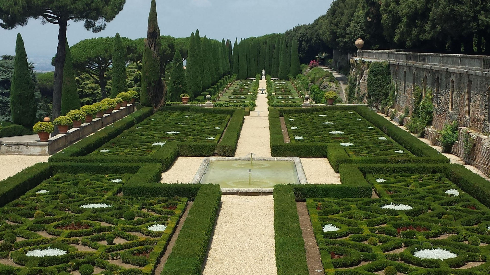 Symmetrical box-hedge gardens around a central pool, with cypresses and an ancient stone wall