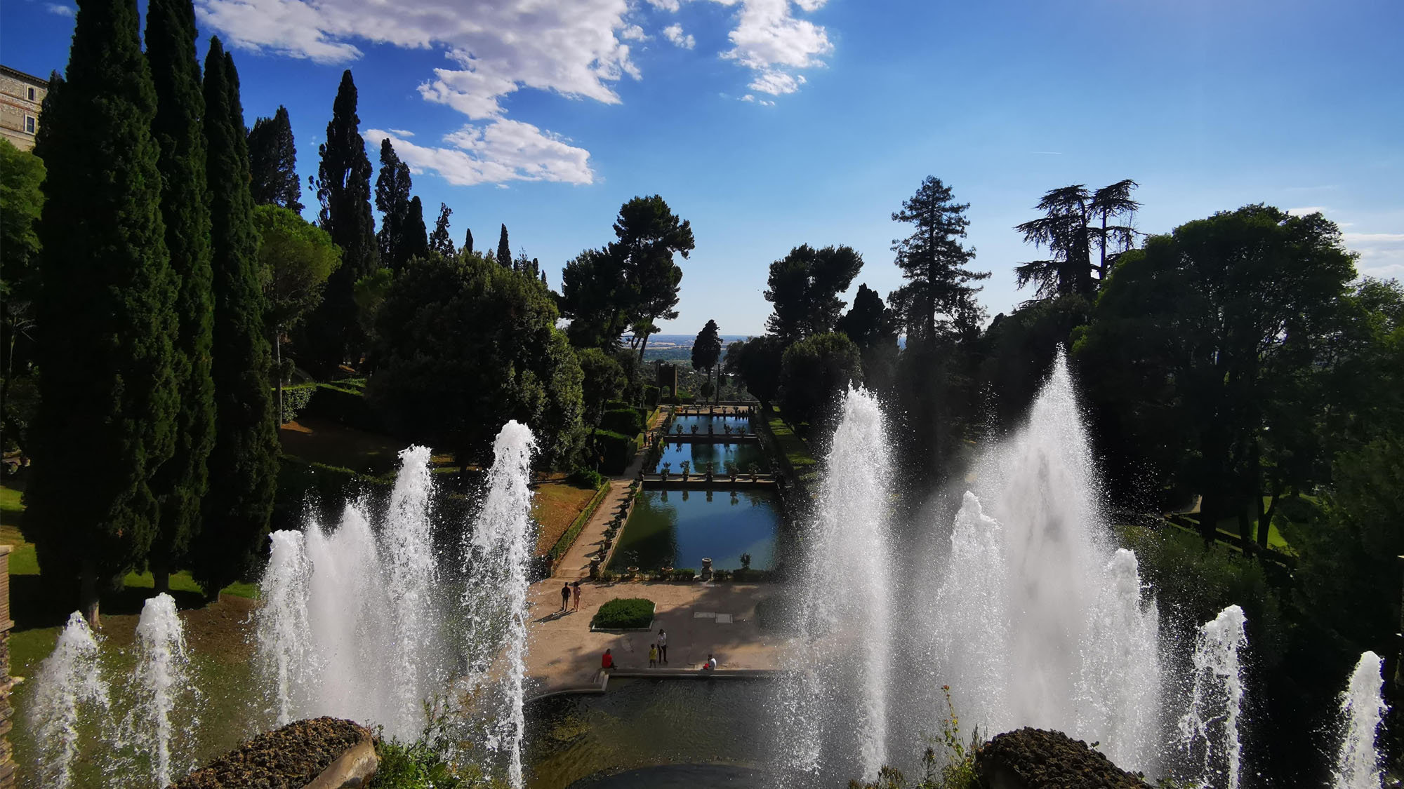 Aerial-style view of garden fountains and cypress trees stretching toward a pool below