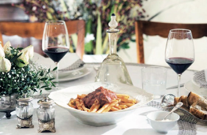 Plate of penne in tomato sauce on a laid table with red wine glasses, bread and white roses