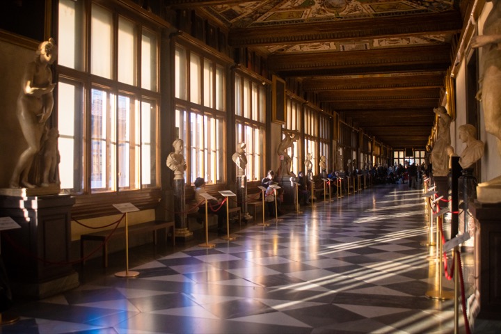 Long Uffizi corridor with marble statues and a chequered floor lit by sunbeams from tall windows