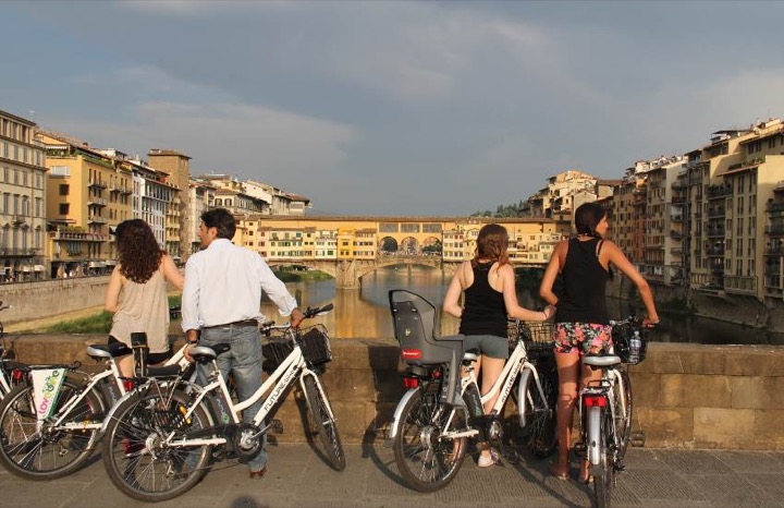 Cyclists on city bikes pausing on the Arno embankment to look towards the Ponte Vecchio at sunset