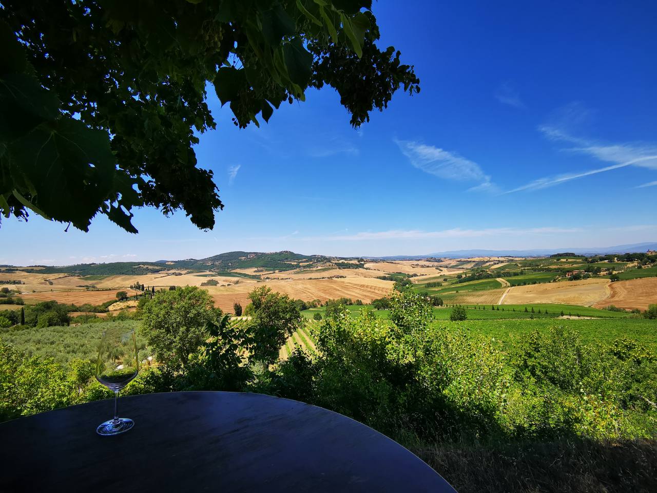 Wine glass on a dark table overlooking rolling Chianti hills, fields and olive groves under a blue sky