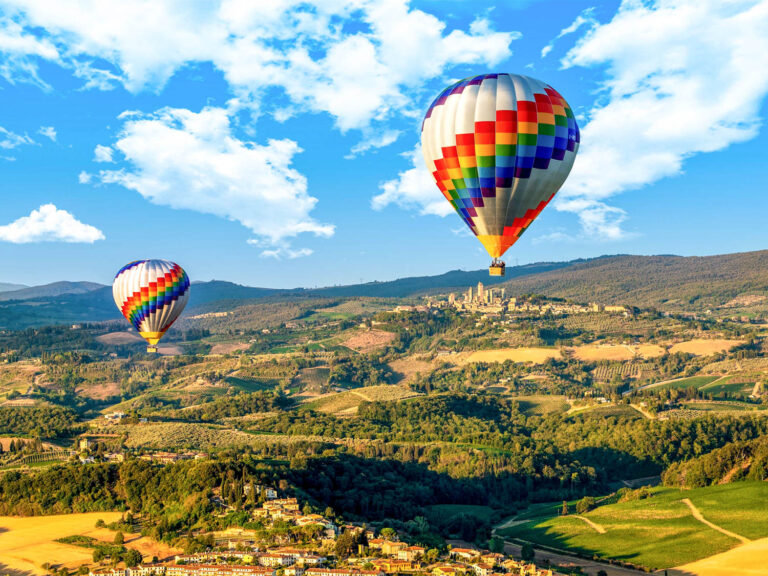 Two rainbow-striped balloons over Tuscan hills with the towers of San Gimignano on the horizon