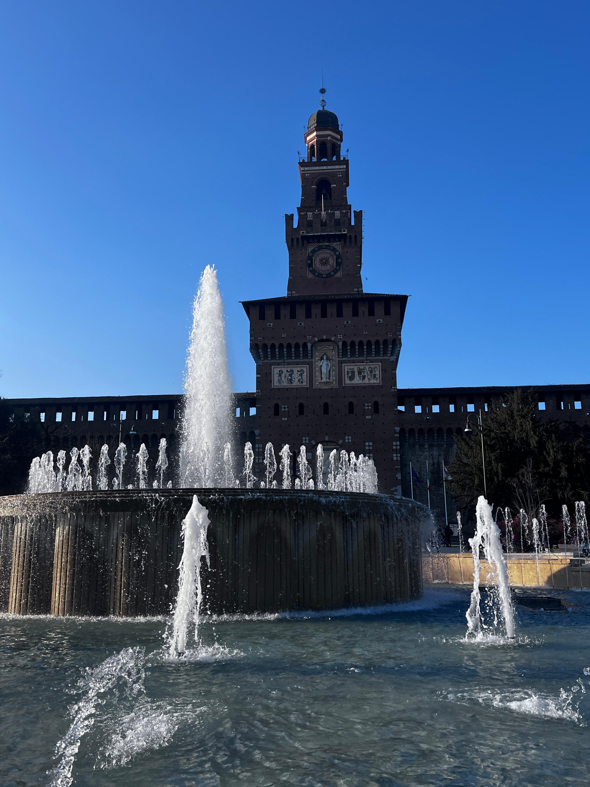 Fountain jets in front of the brick tower of the Sforza Castle in Milan under a clear blue sky