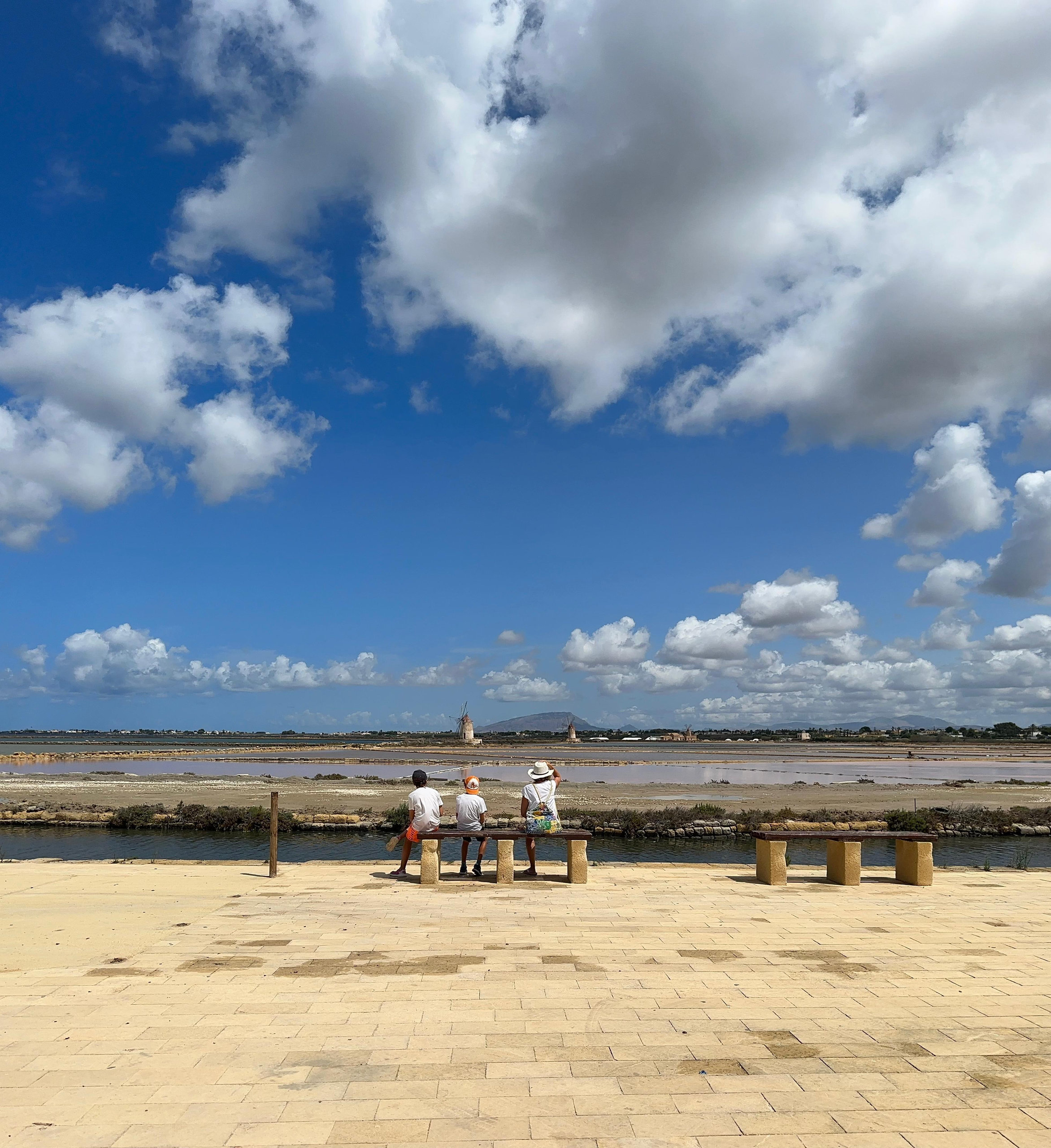 Travellers seated on benches facing salt pans and a distant windmill under a big blue sky