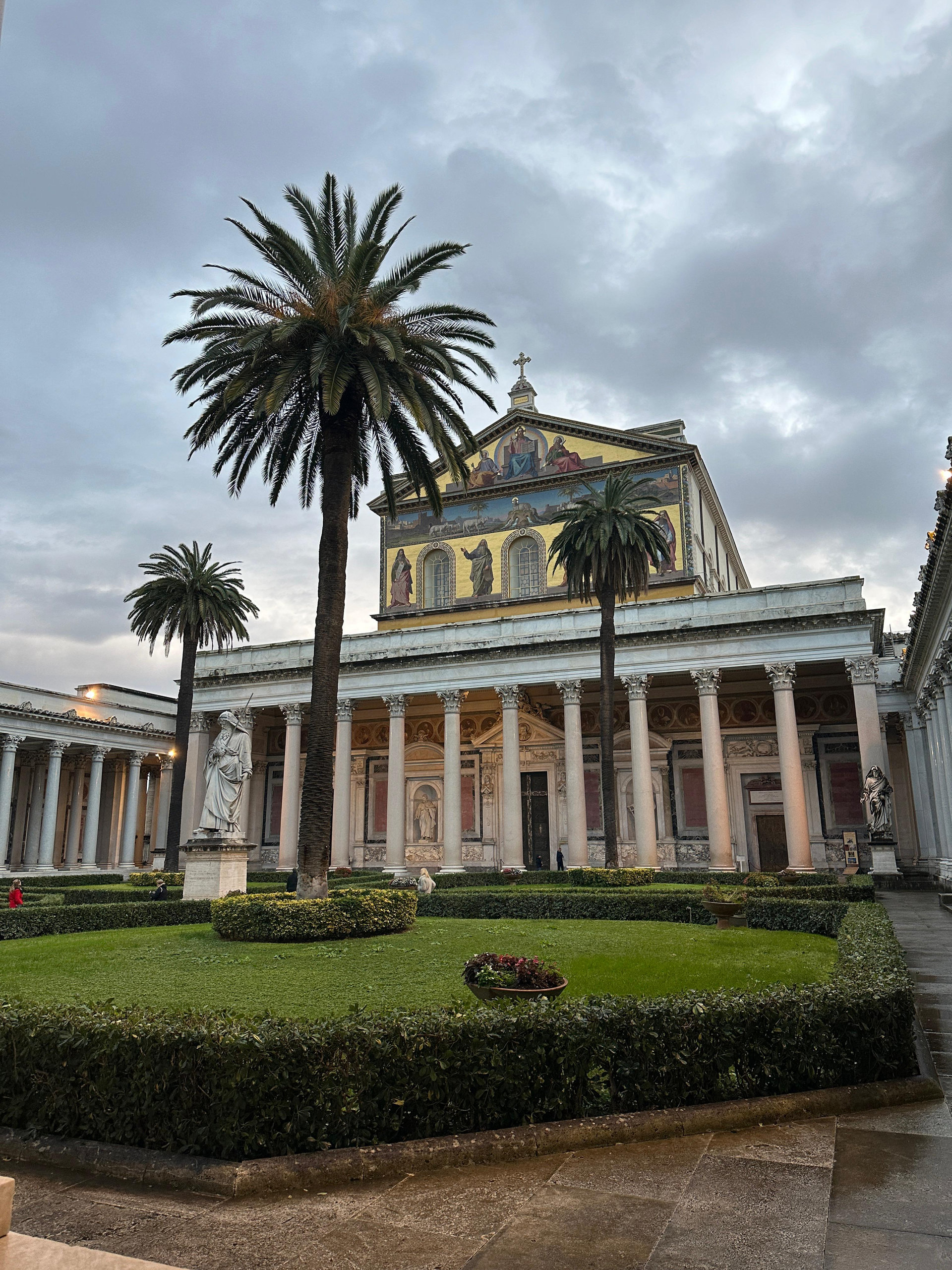 Cloister garden with palms and a statue in front of a mosaic-fronted basilica at dusk