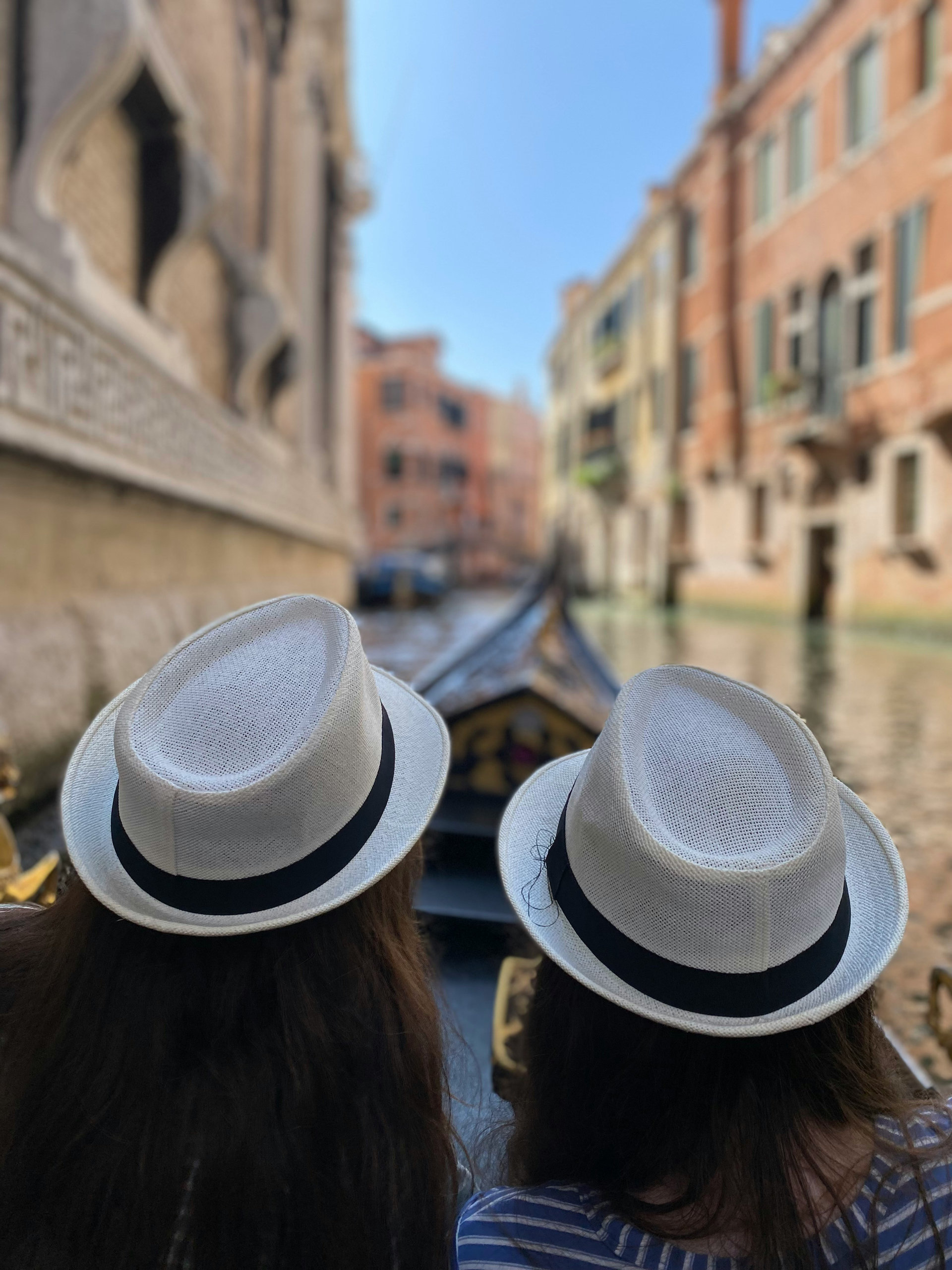 Two travellers in white fedora hats seen from behind on a gondola gliding down a quiet Venetian canal