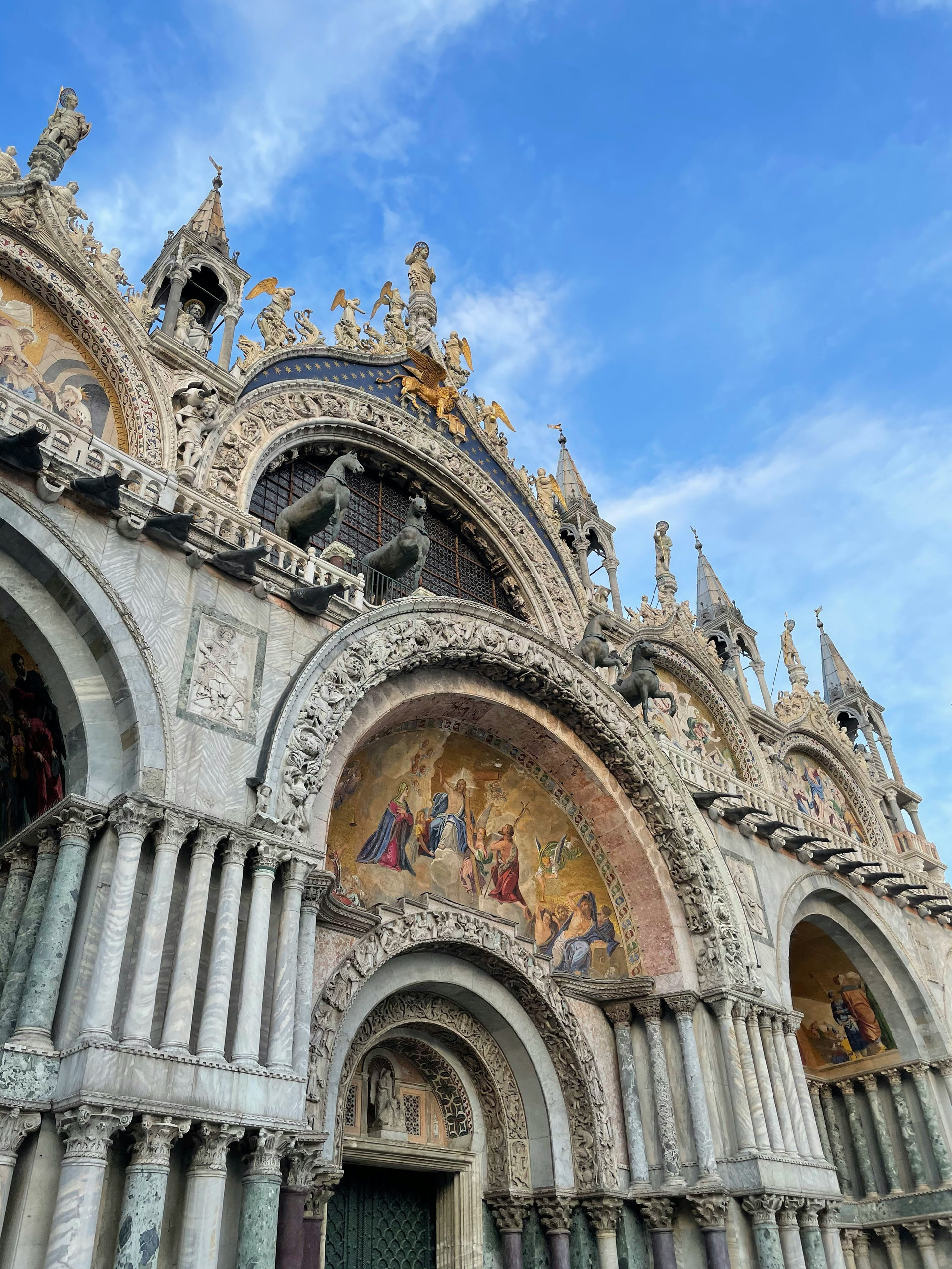 Looking up at St Mark's Basilica, with mosaic lunettes, carved arches and bronze horses against a blue sky
