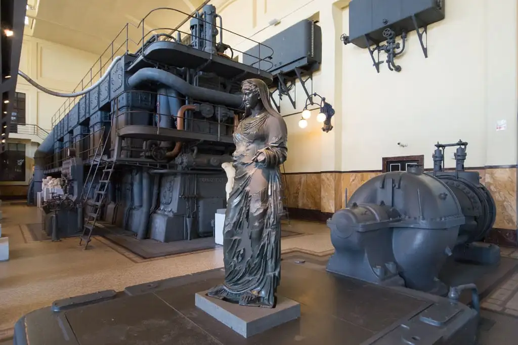 Bronze statue of a draped female figure standing before a large dark diesel engine in the museum hall